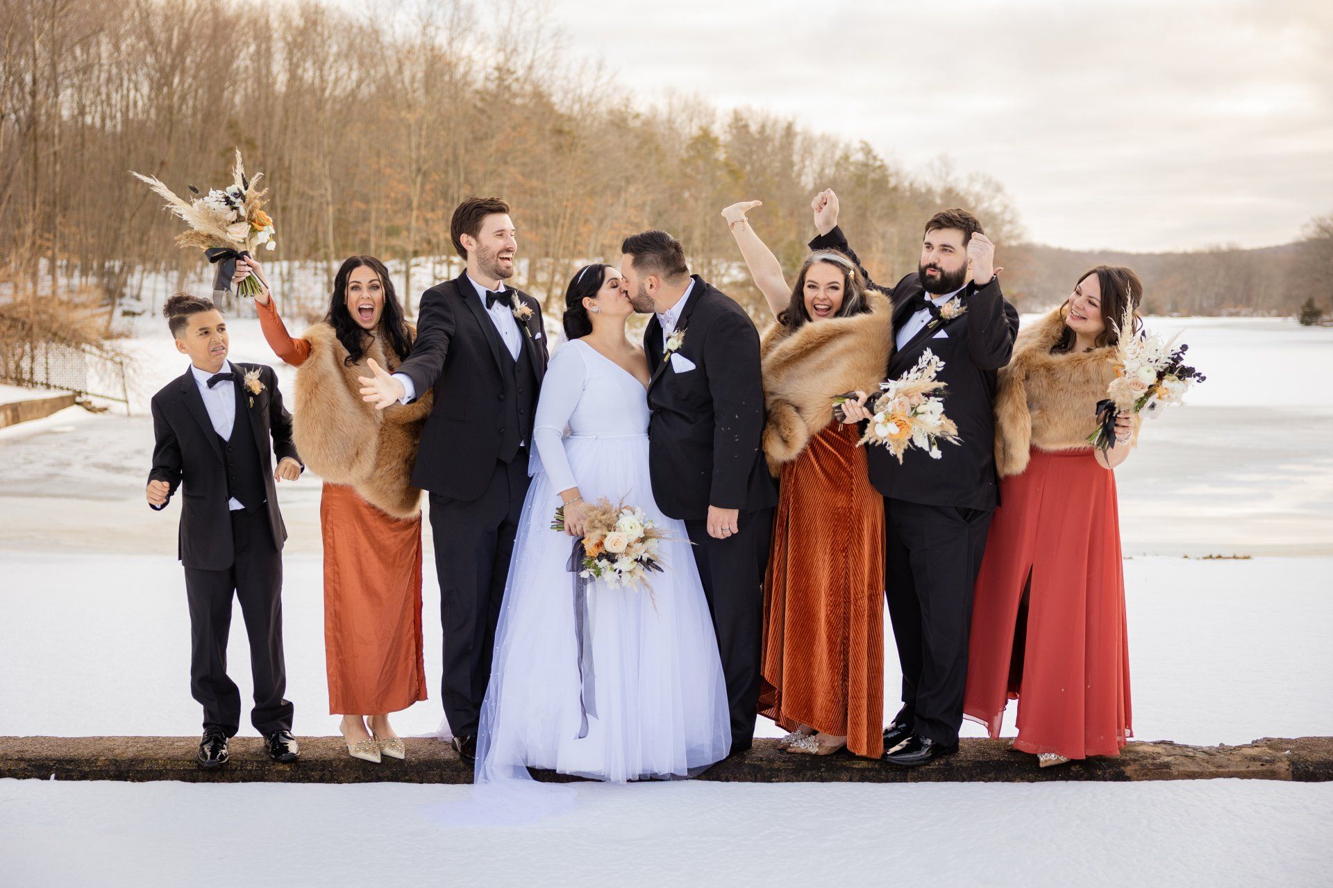 Wedding party posing on a snowy lakeshore with autumn trees, bouquets, and formal attire