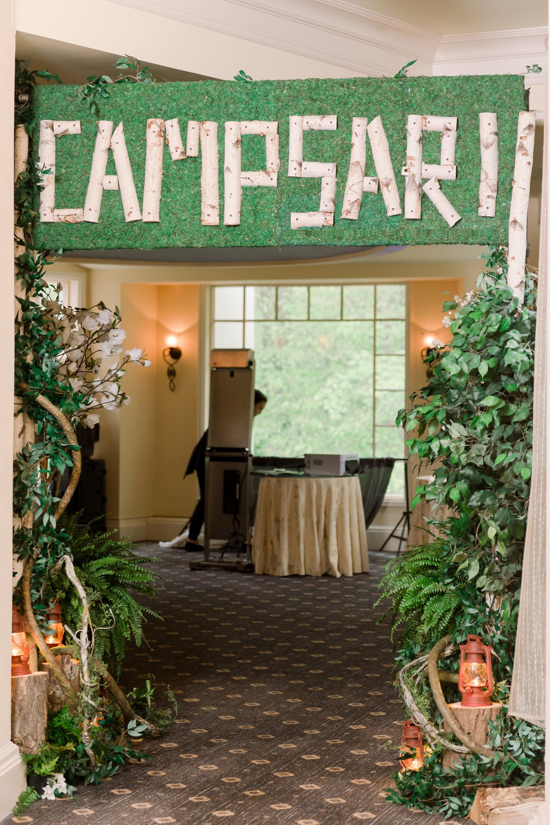 Indoor event entrance with green “CAMPSARI” sign, leafy decor, and a small table in a warmly lit hallway