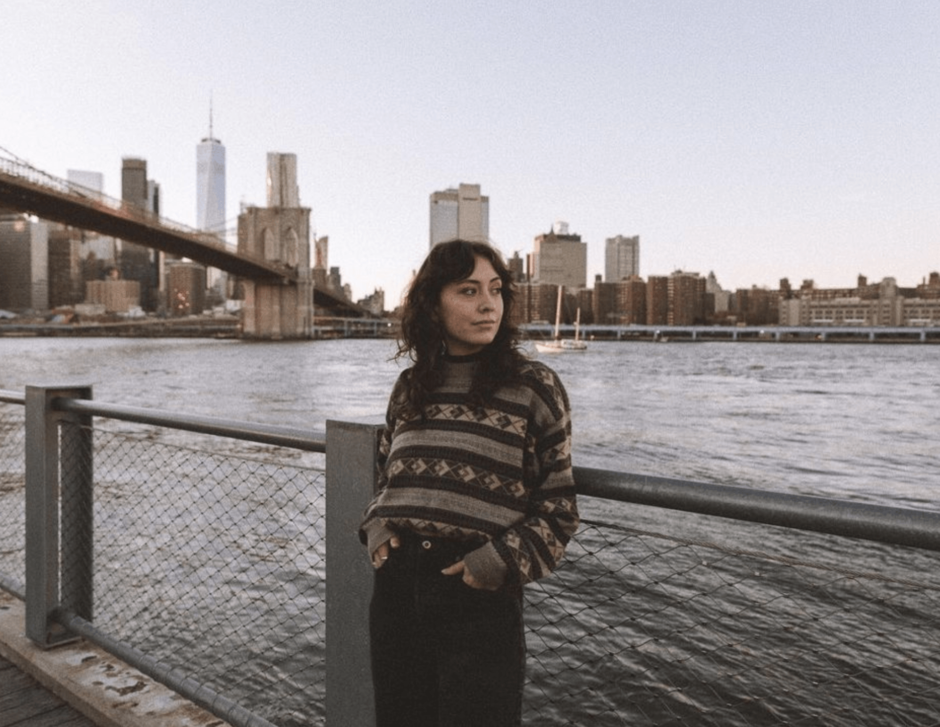 Person in patterned sweater standing by a waterfront railing with a city skyline and bridge in the background