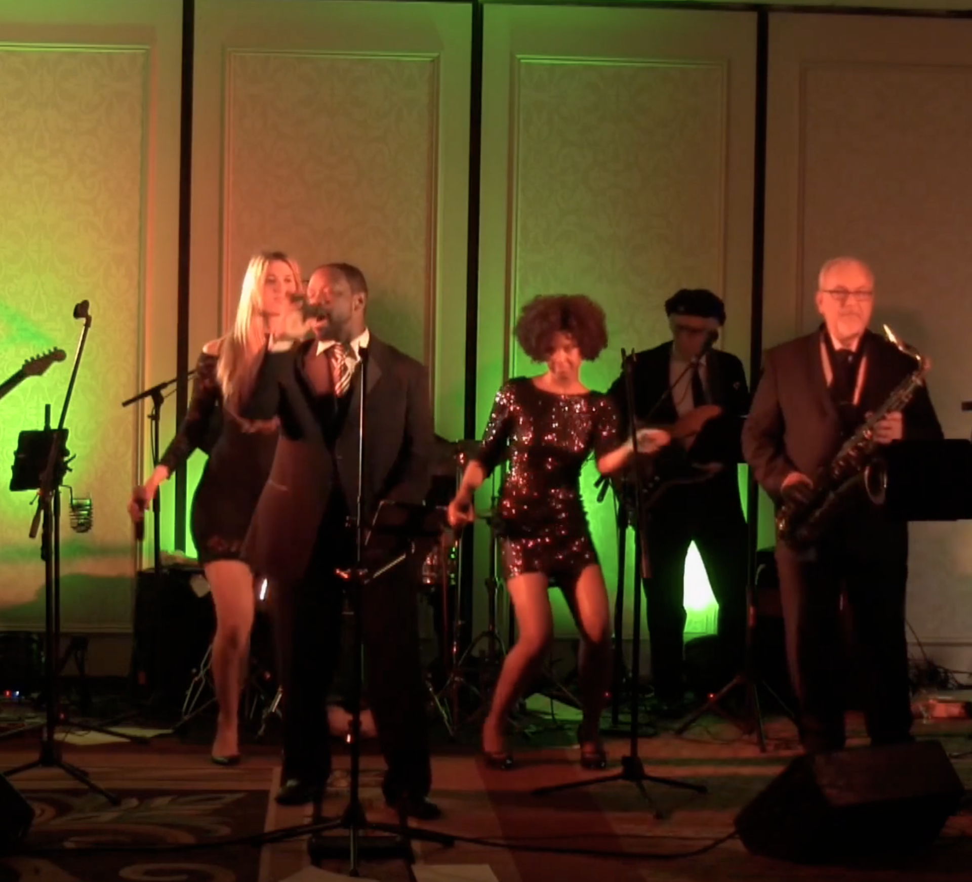 Live band performing on stage under green and warm lights, with singers and saxophonists visible.