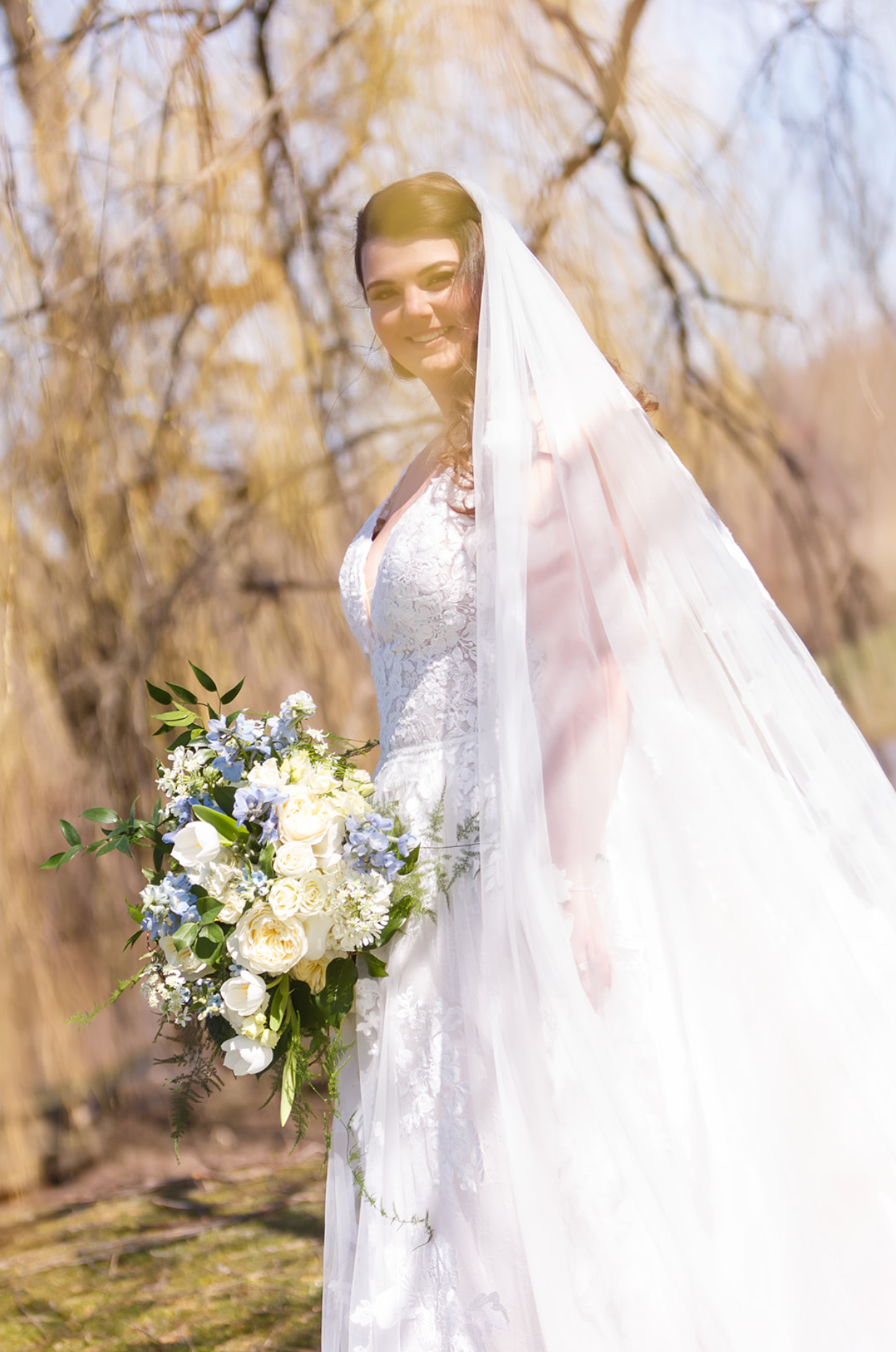 Bride in a white veil holding a bouquet of white and blue flowers outdoors among bare trees