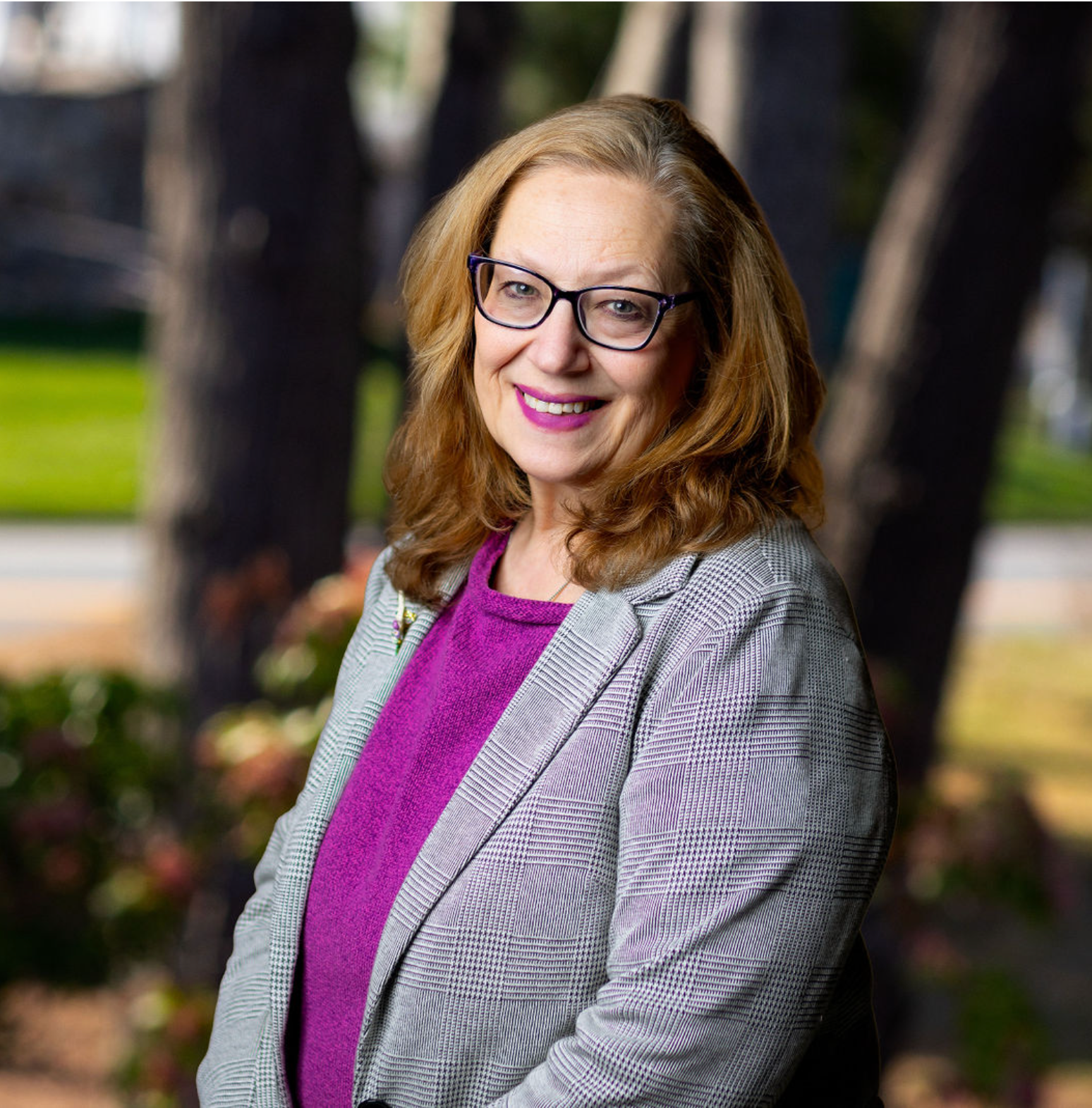 Smiling woman in glasses and a gray blazer standing outdoors in front of blurred trees and flowers