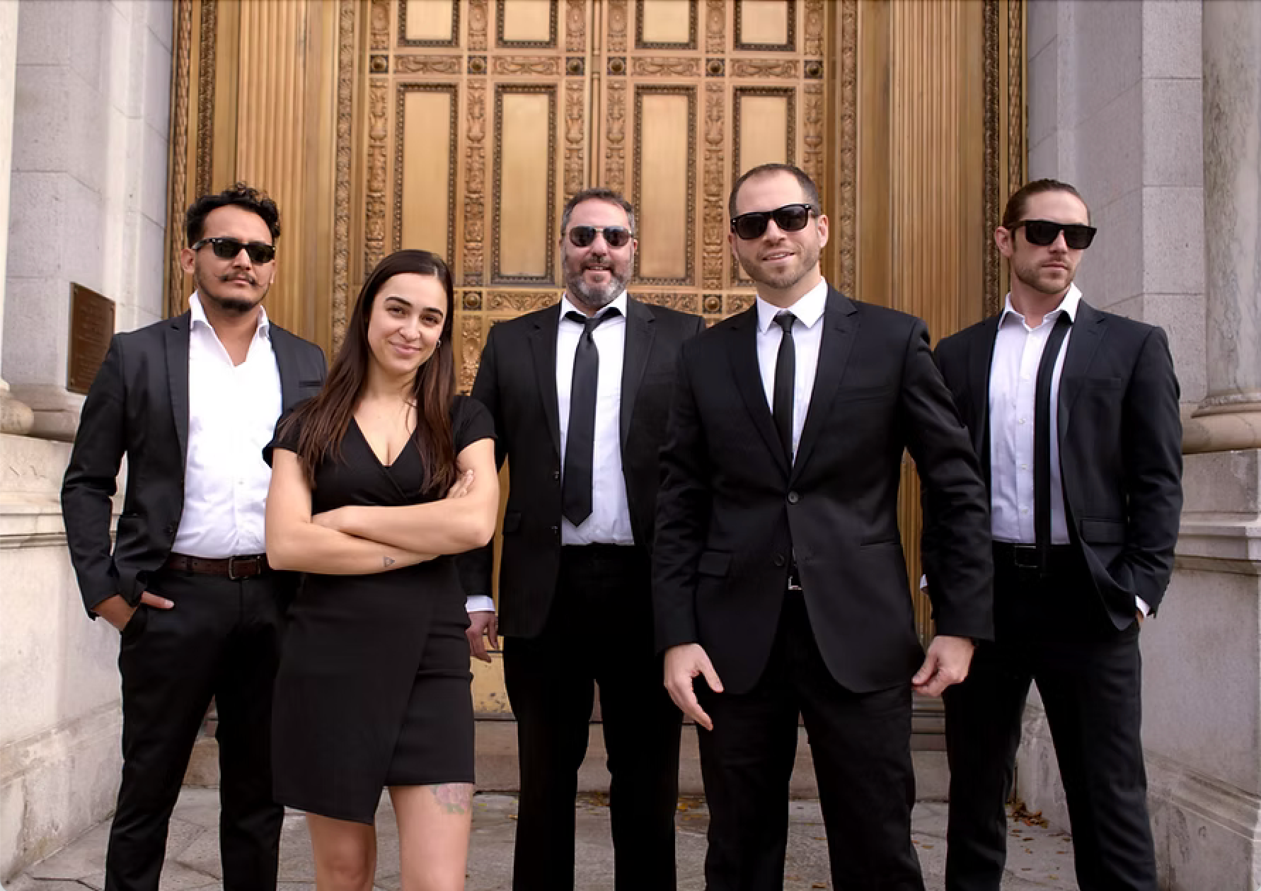 Five people in black formalwear and sunglasses stand outside a stone building, posing together.