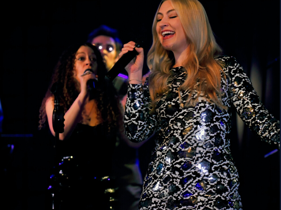 Three musicians performing onstage under blue lighting, with microphones and a smiling singer in the foreground