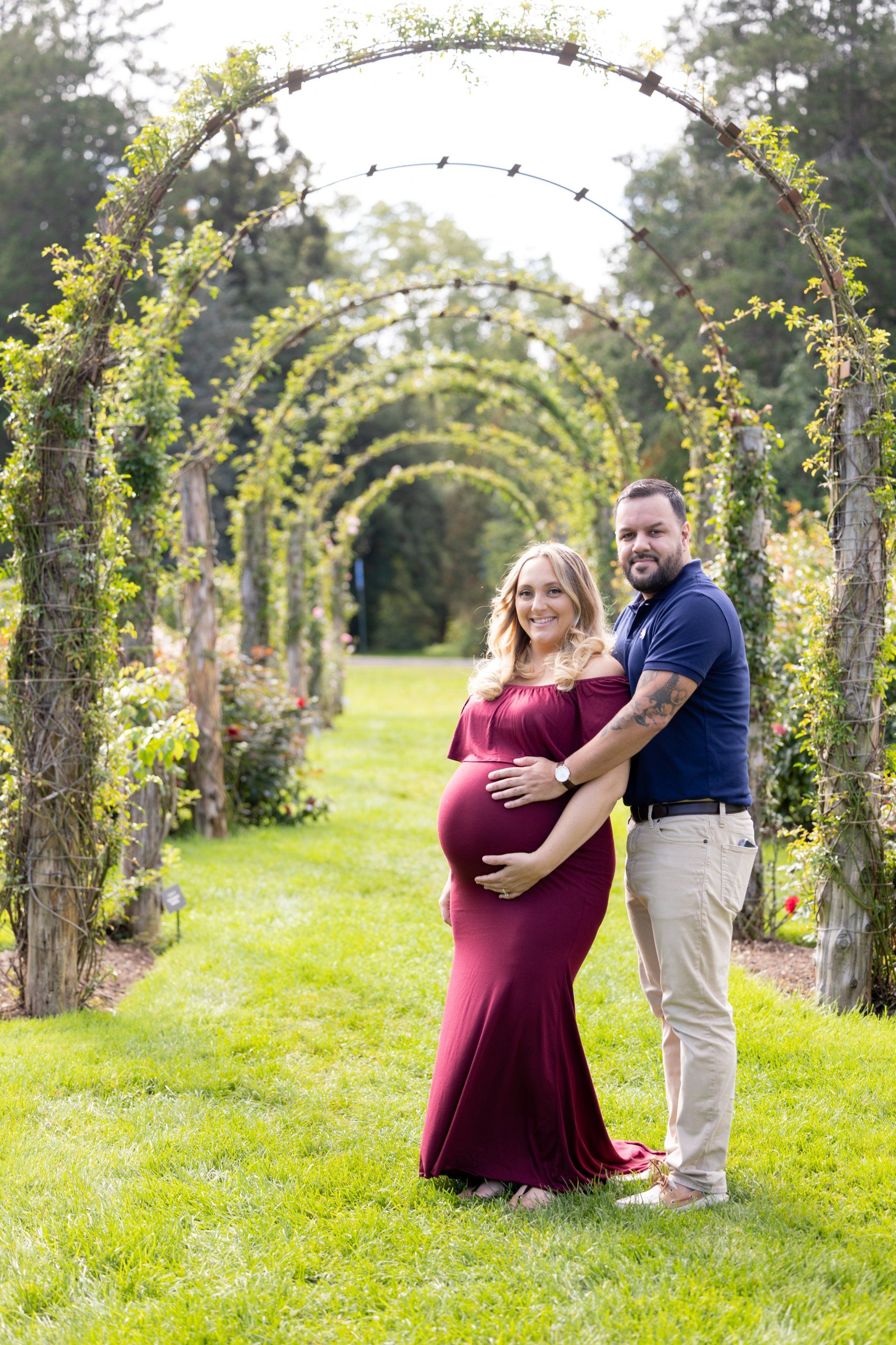 Pregnant couple smiling under garden arches, with the partner holding her belly
