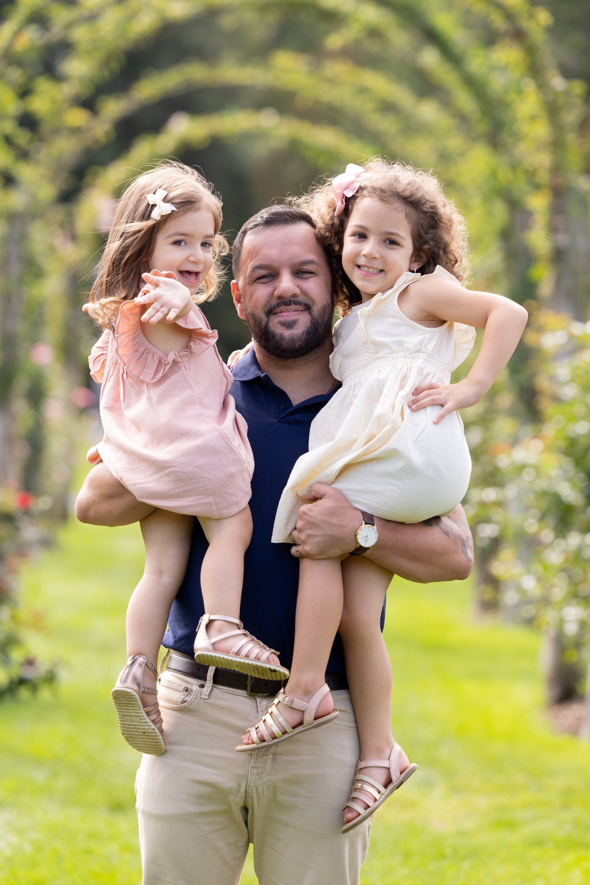 Man in a navy shirt holding two smiling children in pink and white dresses outdoors on grass.