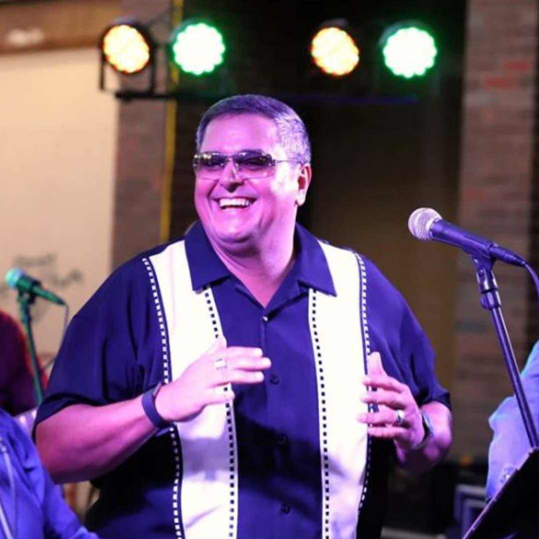 Smiling singer in sunglasses holding a microphone onstage under colorful lights
