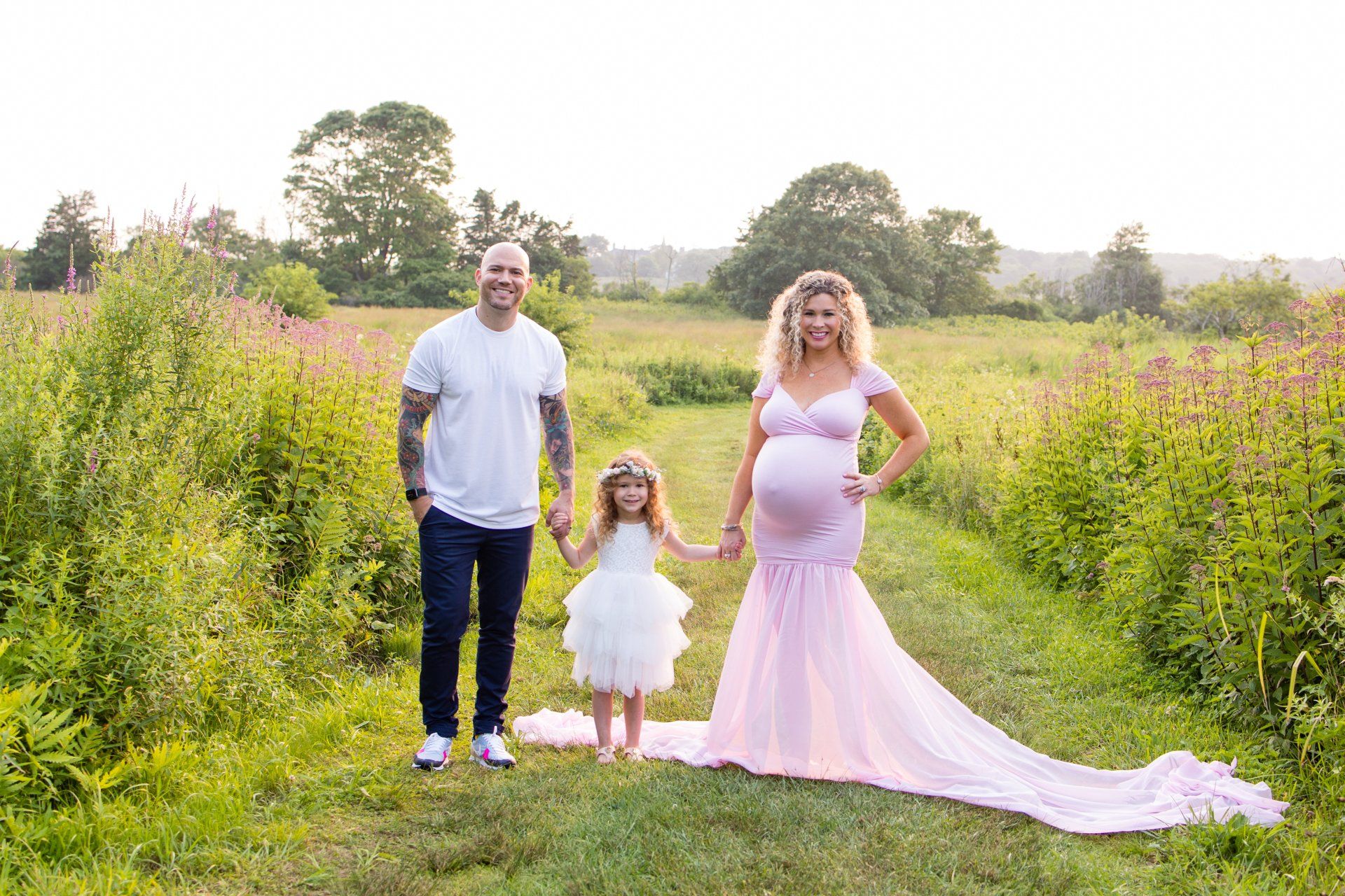Three people posing in a grassy field, with a woman in a pink dress and a long flowing train.