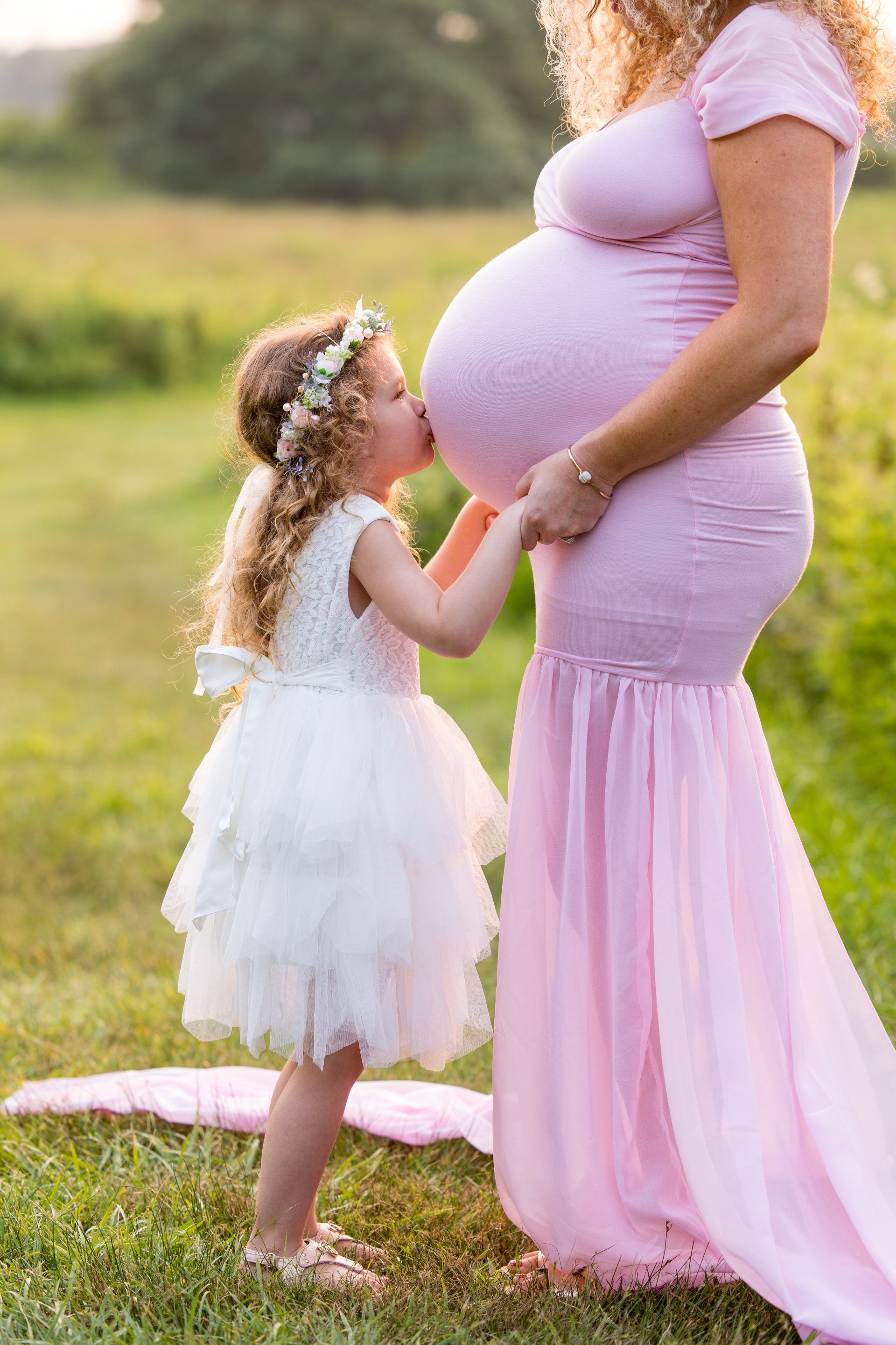 Pregnant person in a pink dress with a young child in a white dress touching their belly outdoors