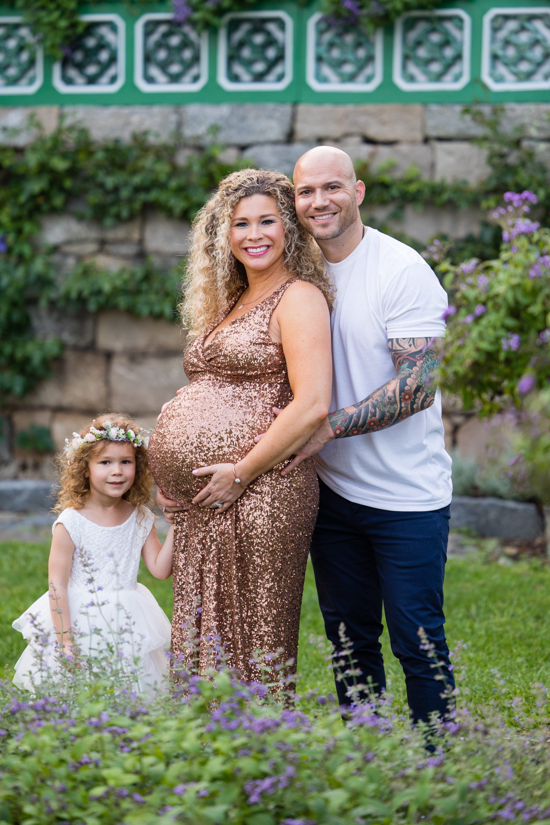 Family posing in a garden, with a pregnant woman in a gold dress and a child in white beside a man in a white shirt.