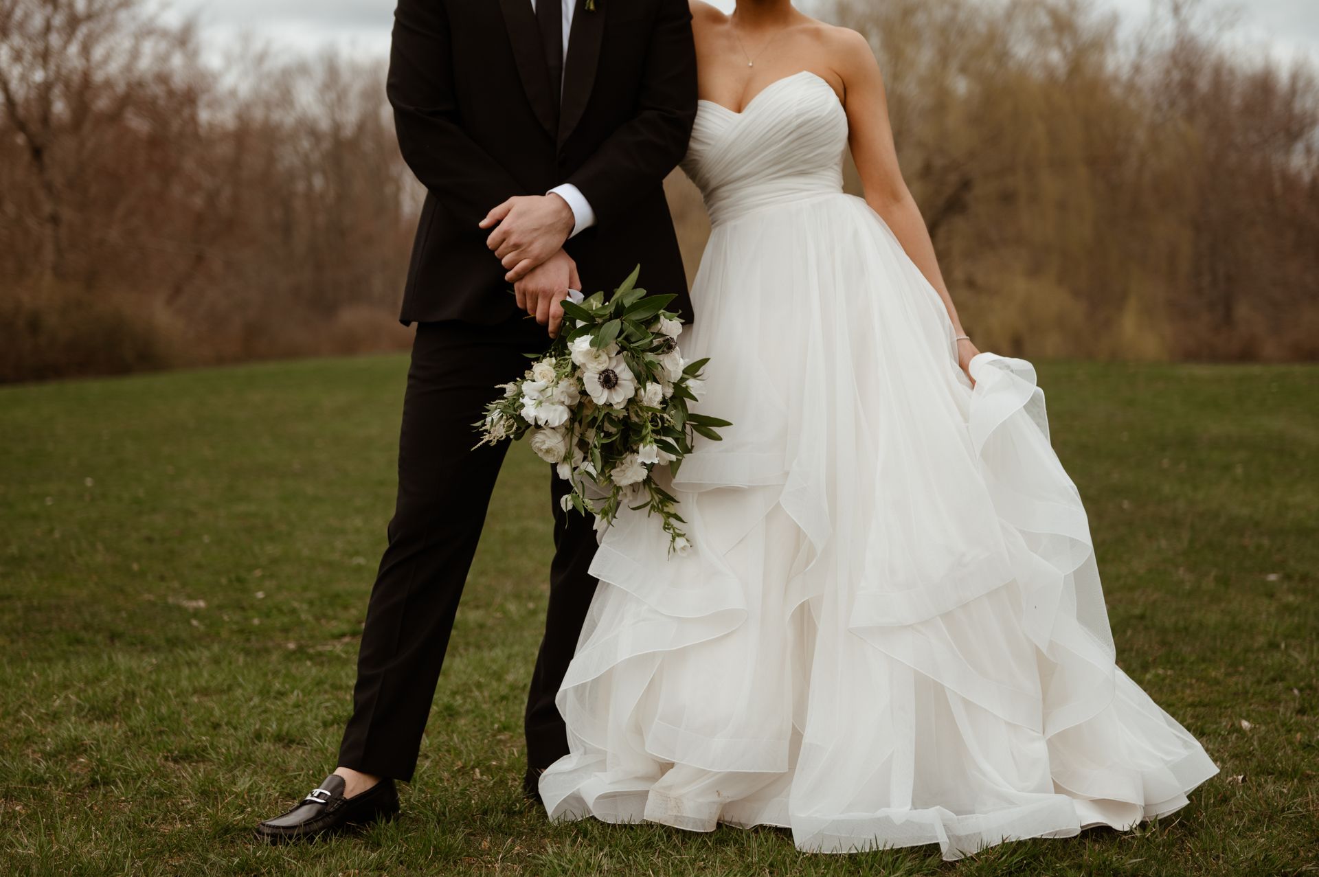 Bride and groom standing outdoors, holding a white floral bouquet in a grassy field