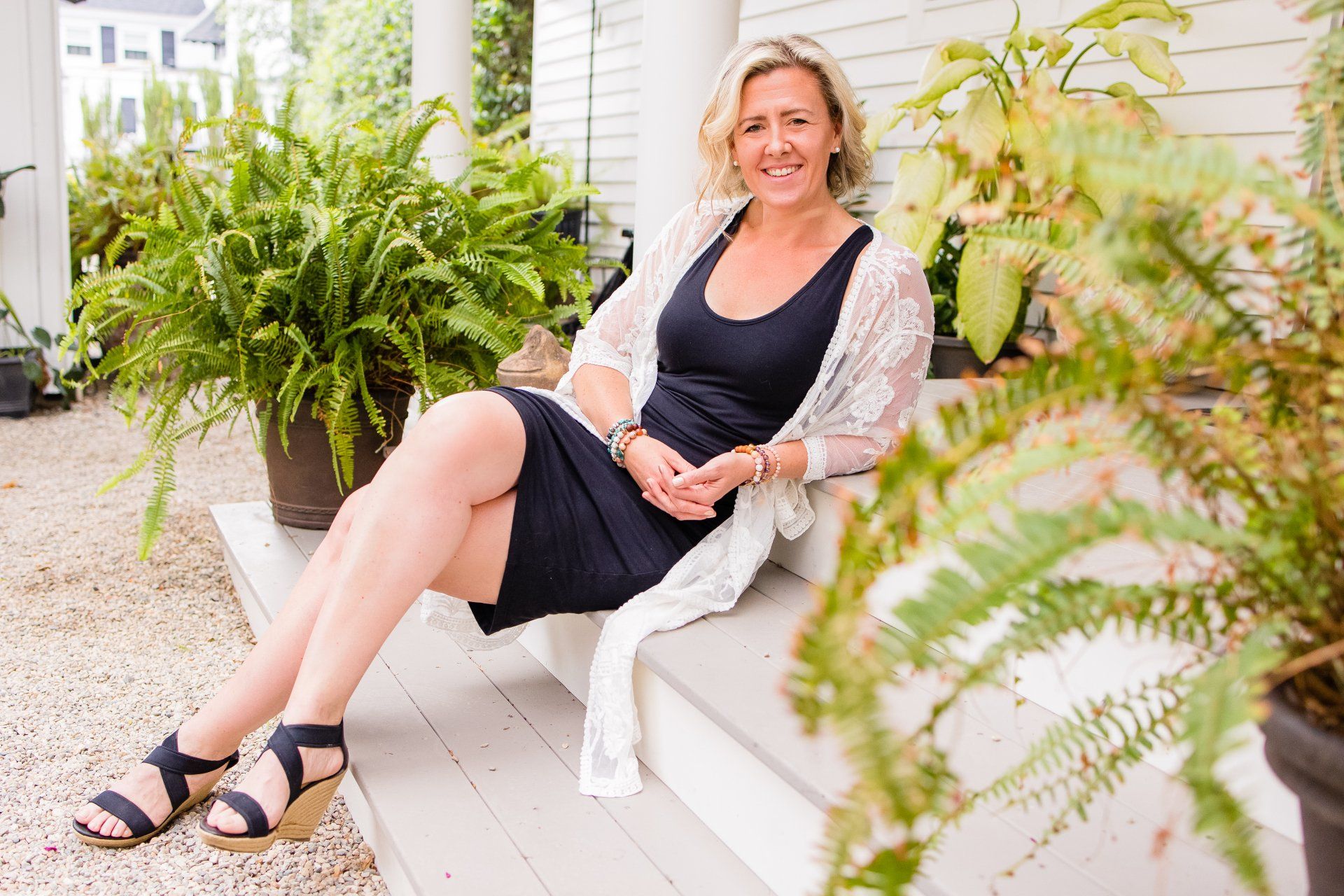 Woman sitting on a porch bench with potted ferns, smiling in a black dress and light cardigan