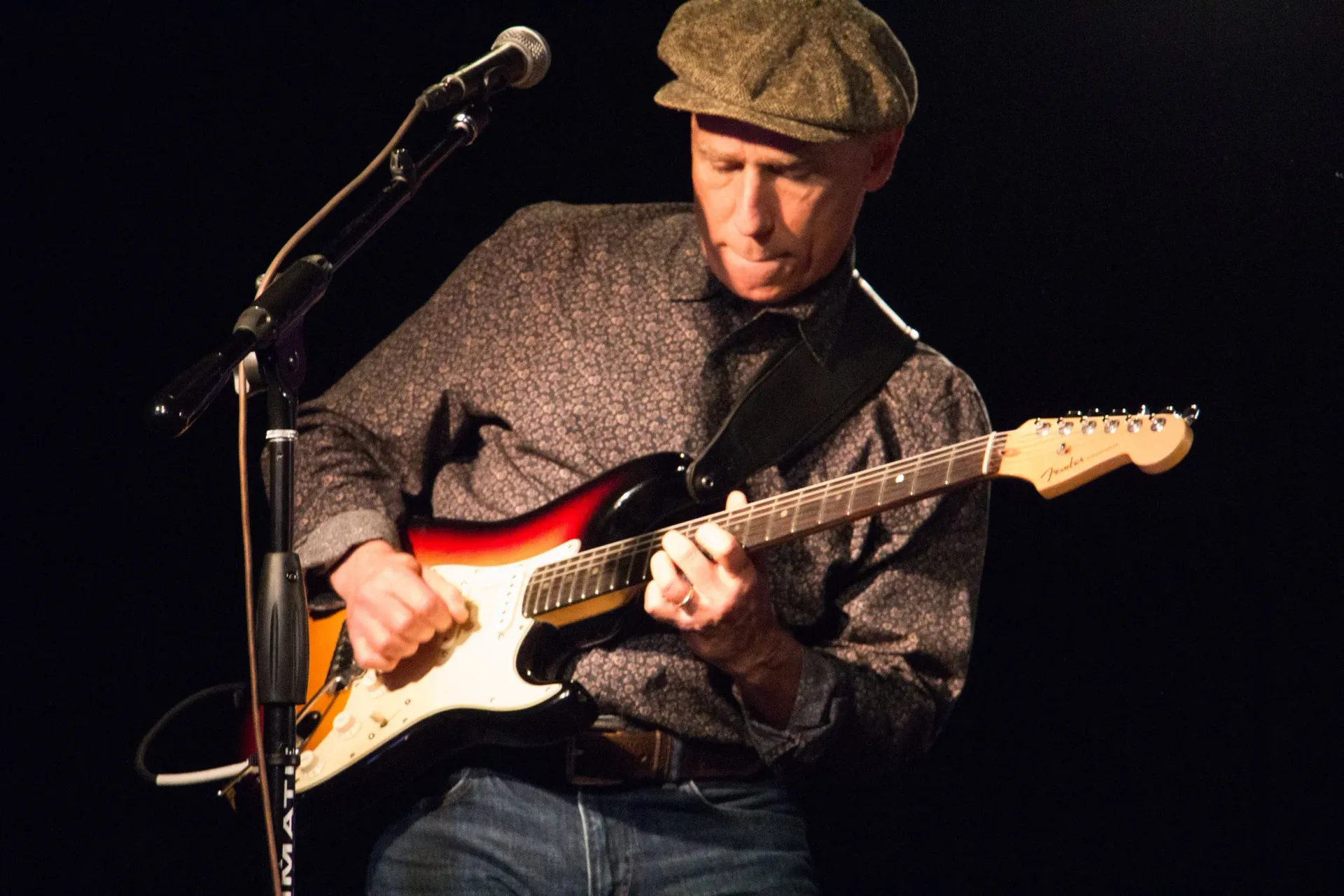 Musician in a flat cap playing electric guitar on a dimly lit stage beside a microphone stand