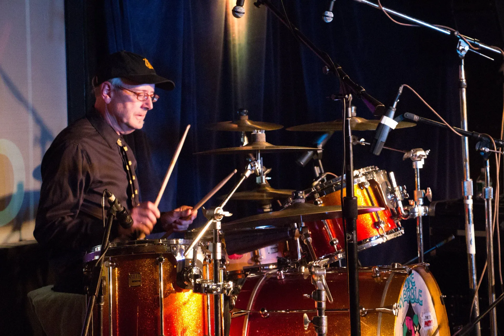 Drummer playing a red drum kit on stage under warm lights