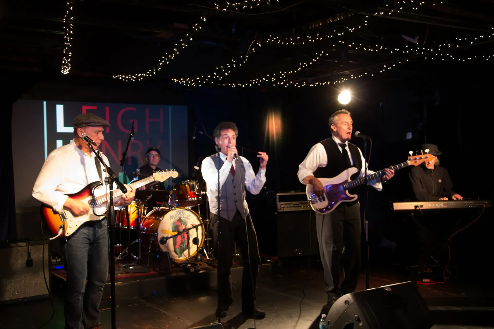 Live band performing on a small stage with guitars, drums, and microphone under string lights