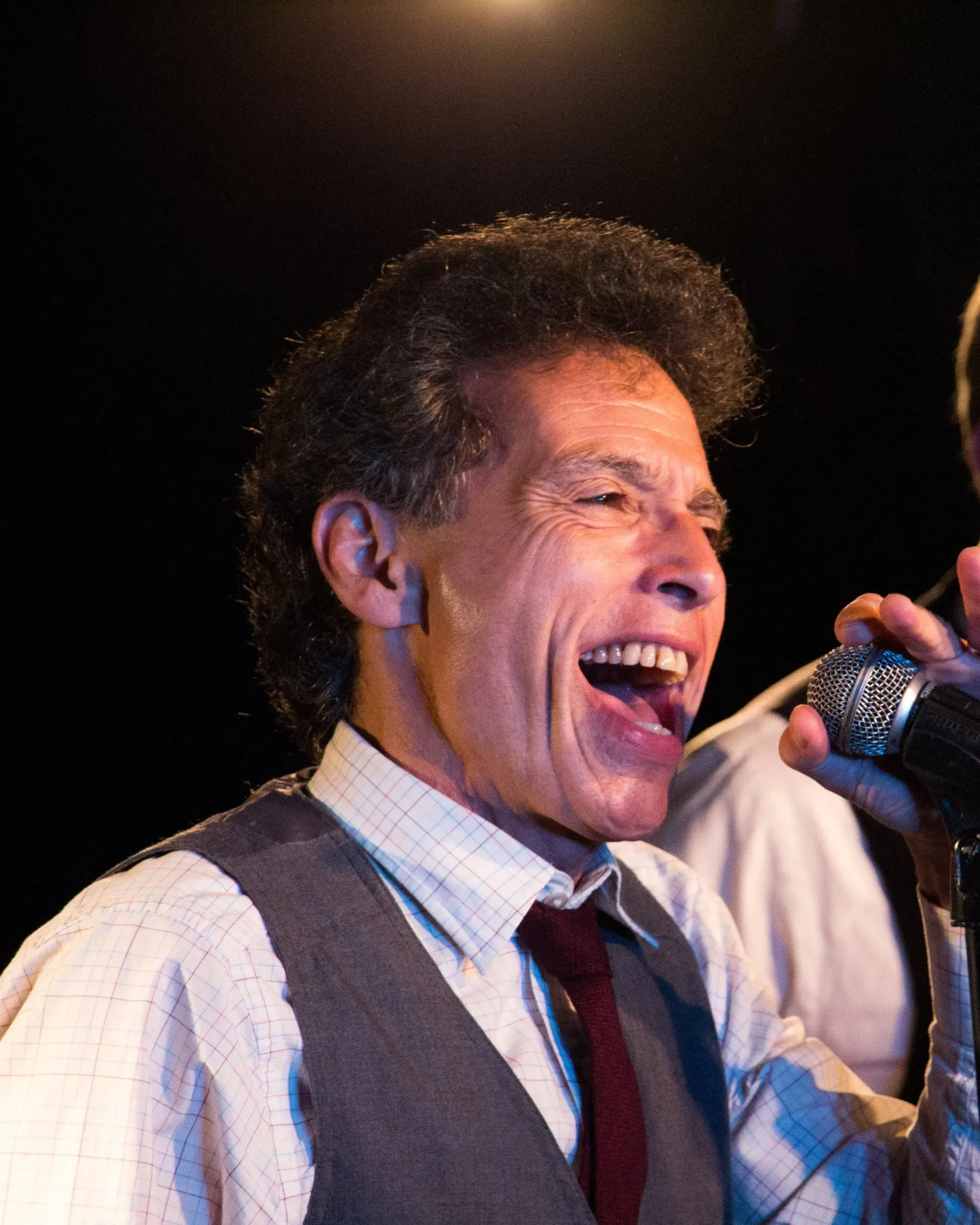 Man singing into a microphone on stage, wearing a vest and tie, with a lively expression
