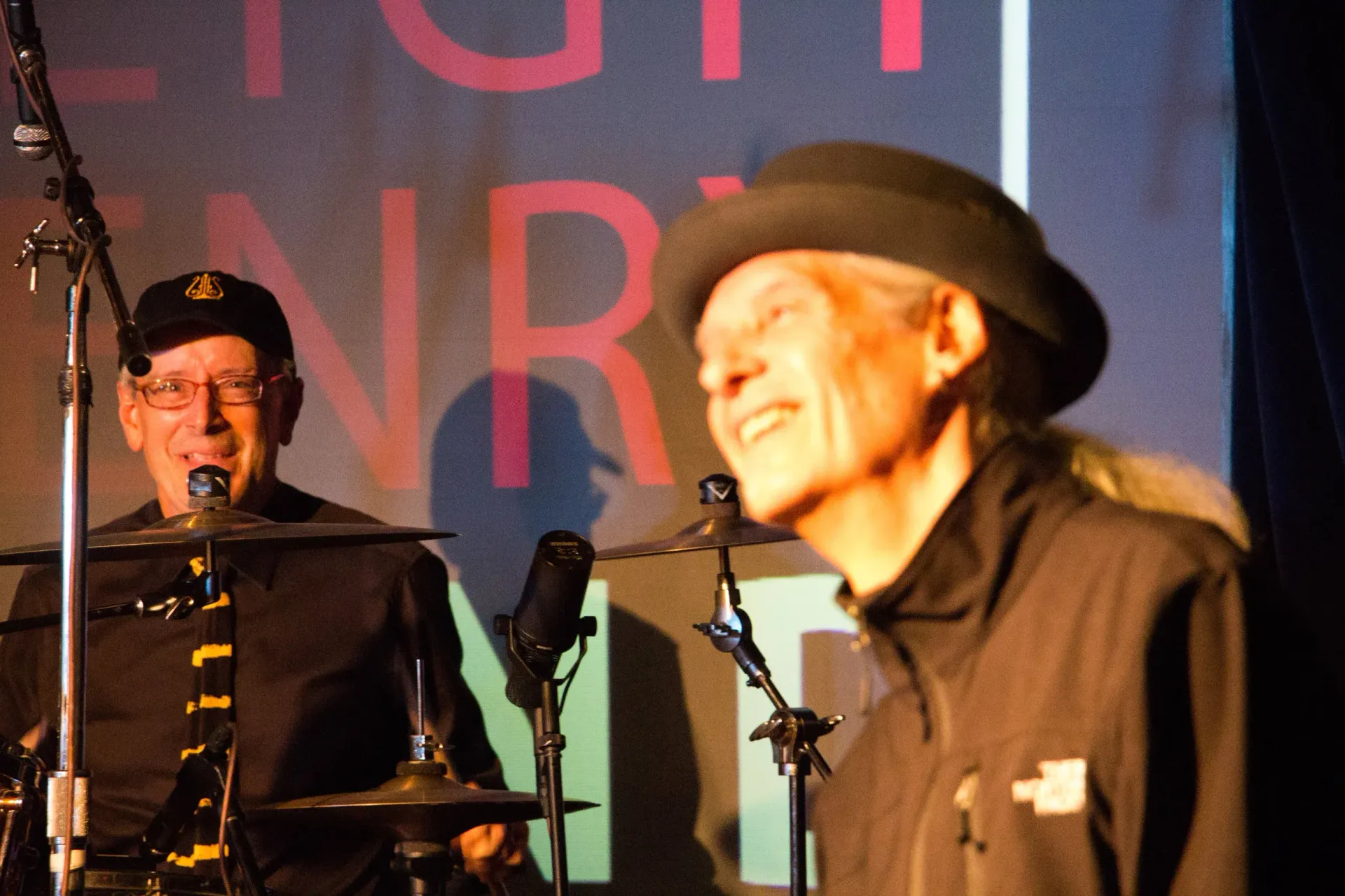 Two musicians on stage, one smiling in a black hat and one at the drum kit, with microphones and red lettering behind them
