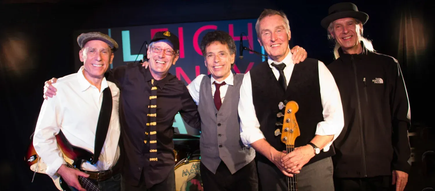 Five men posing onstage, smiling, with one holding a guitar, under colorful lights.