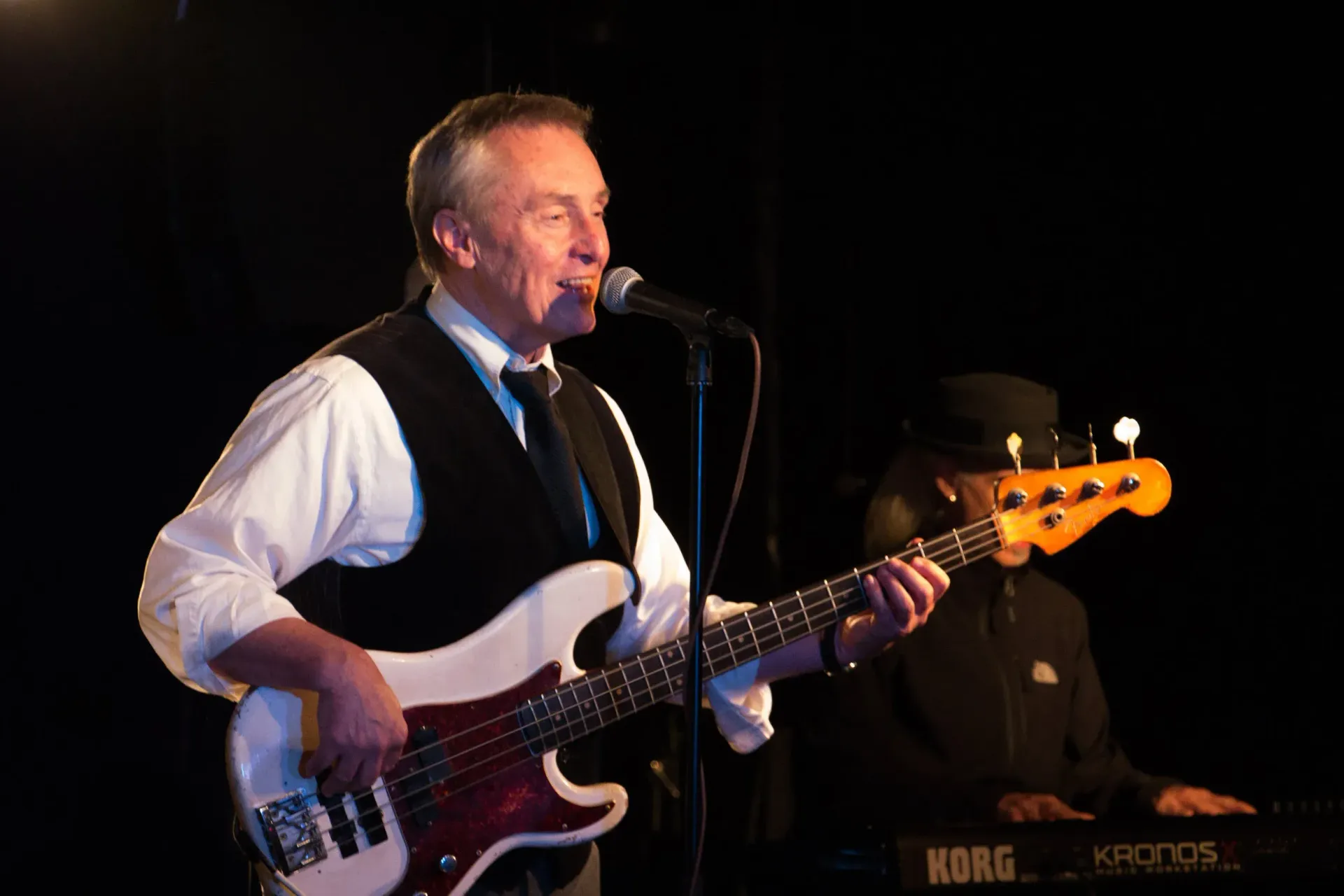 Musician singing into a microphone while playing a bass guitar on stage, with a keyboard player in the background