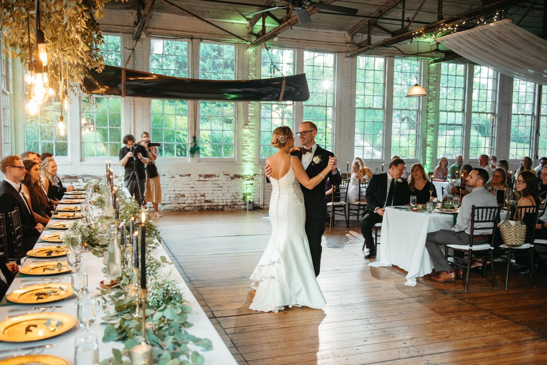 Bride and groom dancing at a rustic indoor wedding reception with guests seated at long tables