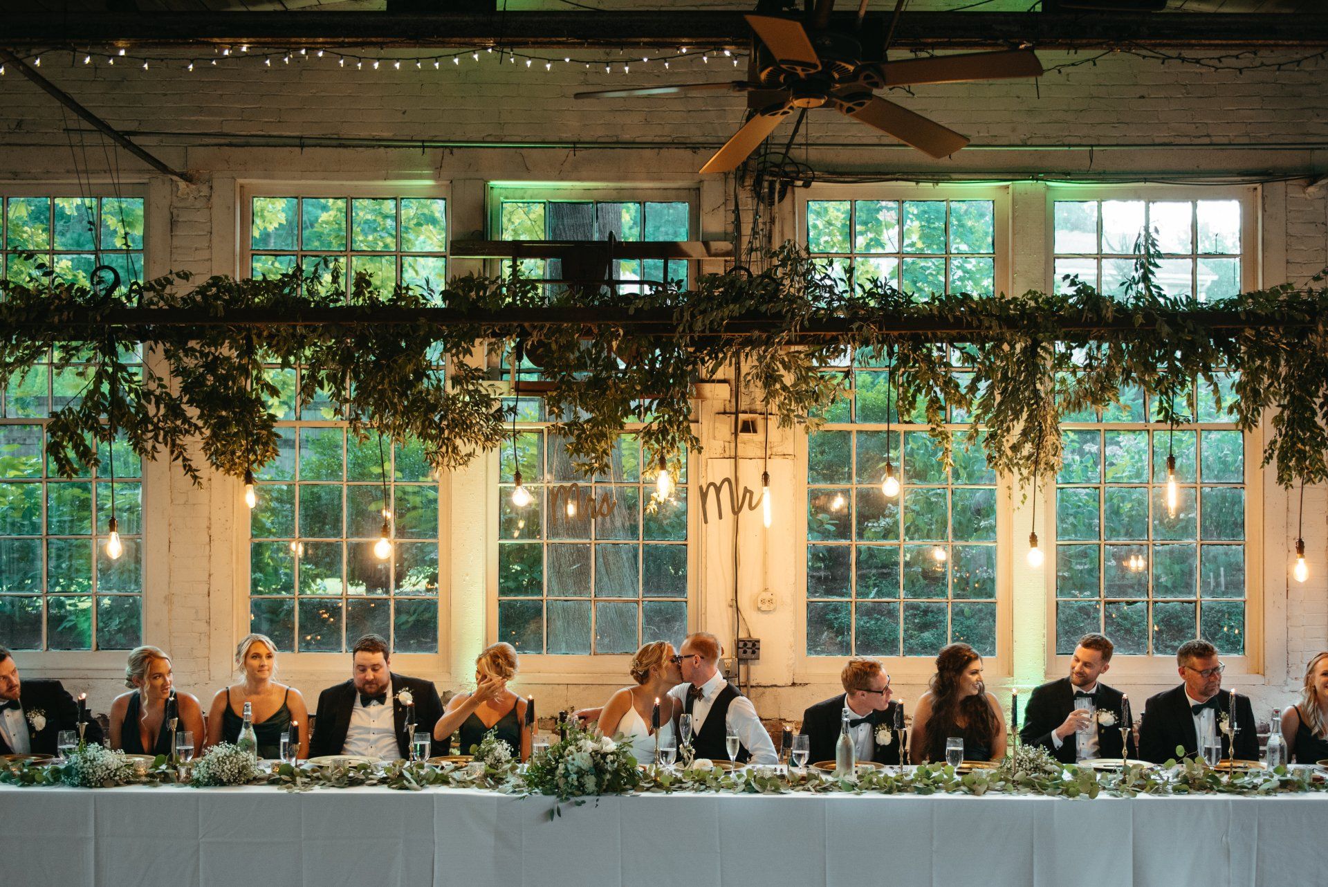 Long banquet table with seated guests under hanging greenery in a bright glass-walled hall