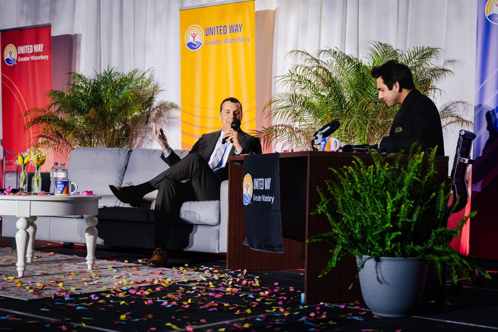 A man sitting on a couch talking to the event host who is sitting at a desk. 