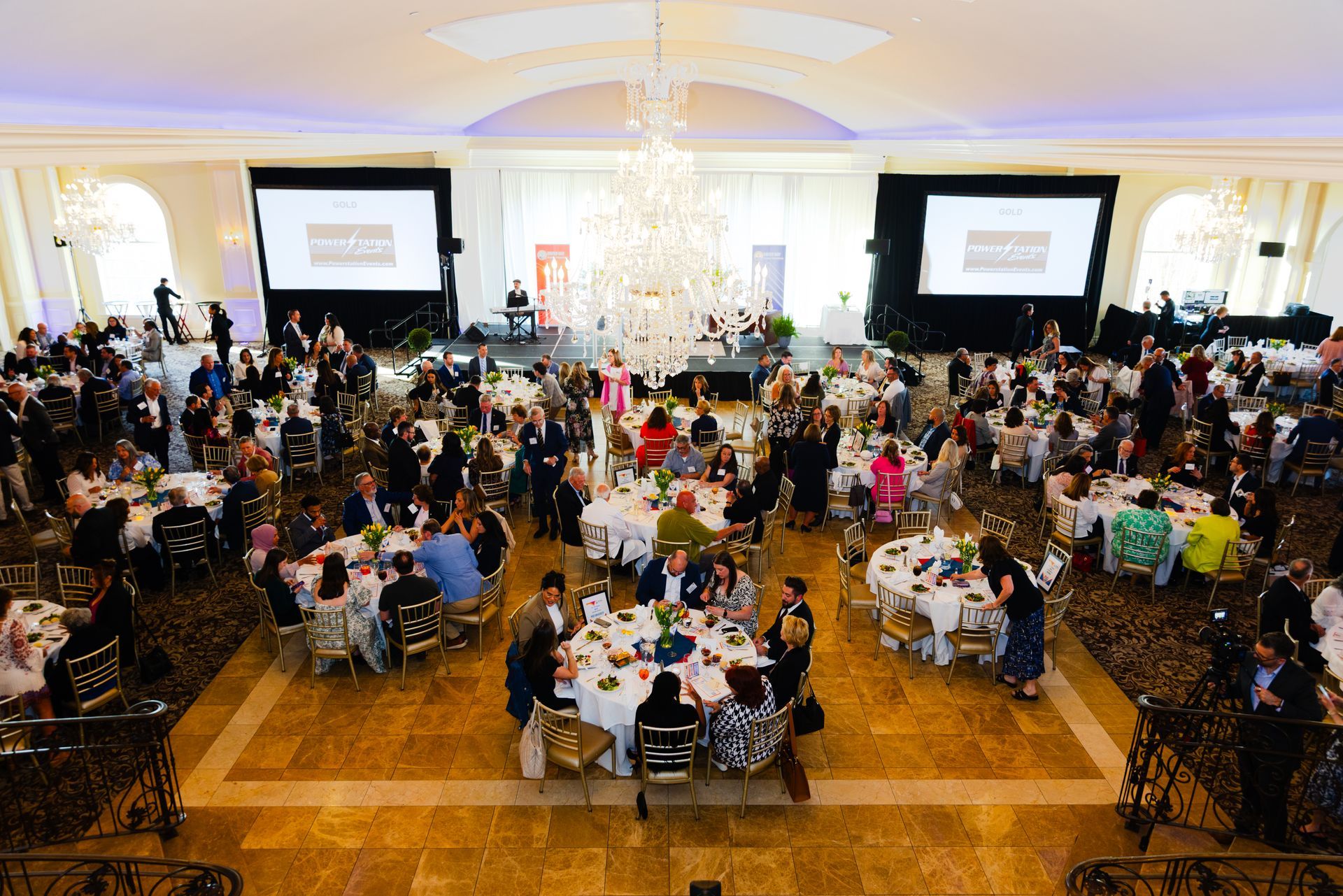 Large banquet hall filled with seated guests at round tables beneath chandeliers, with two projection screens at the front