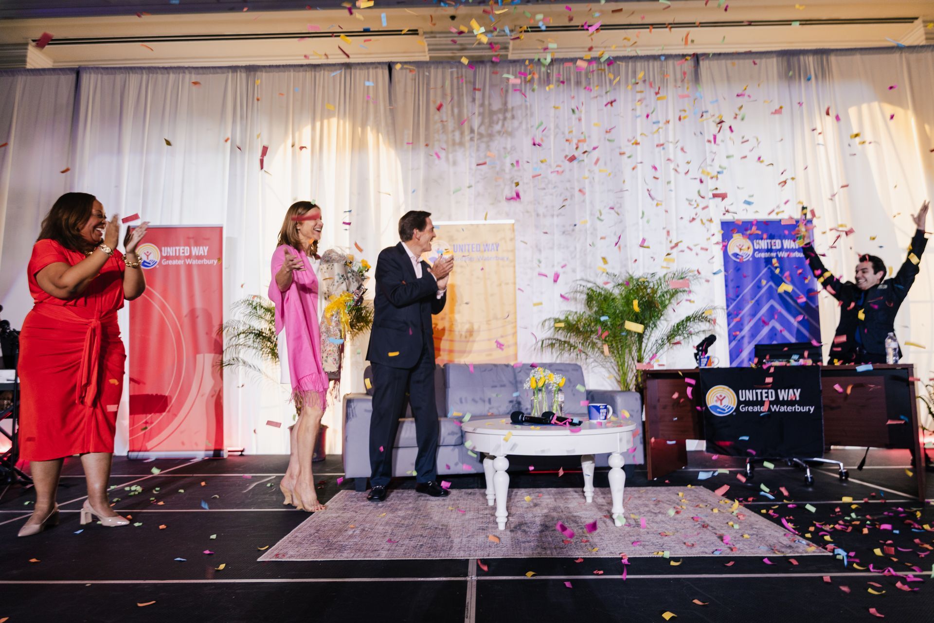 Four people on a stage with confetti falling, one speaking at a podium during an event.