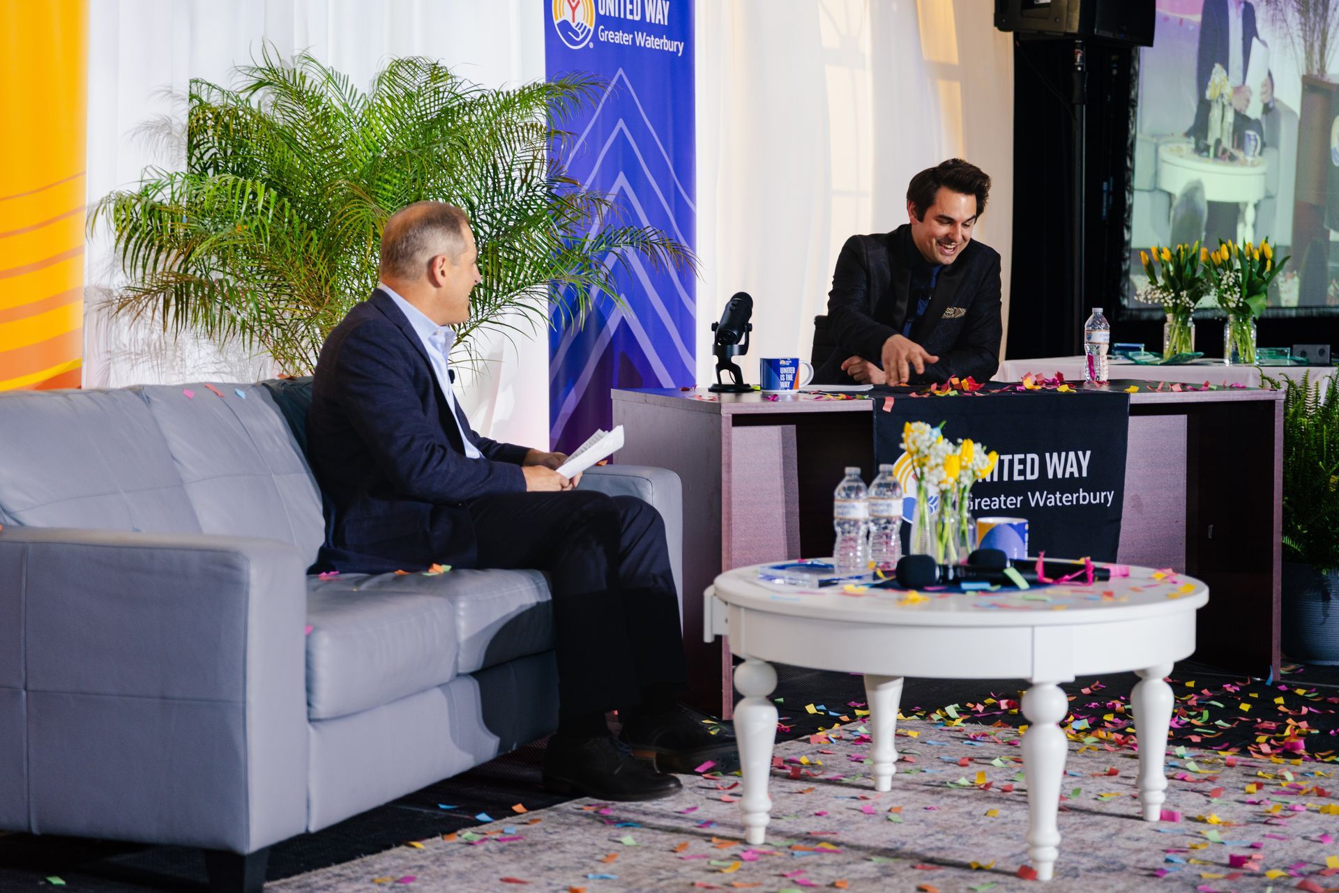 Two men seated in a bright lobby lounge, one speaking at a desk during an interview or discussion.