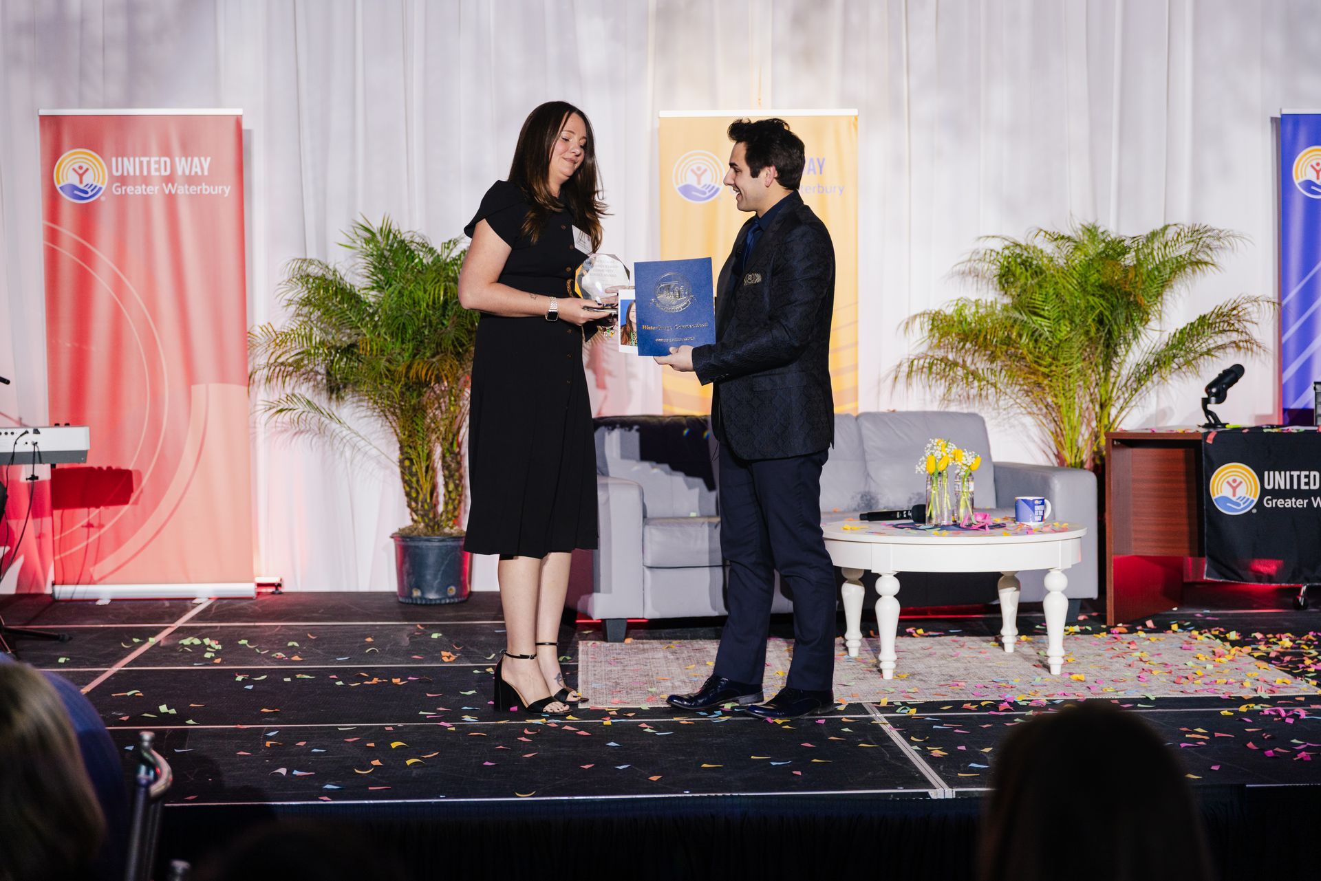 Two people onstage exchange a certificate amid confetti at a formal ceremony.
