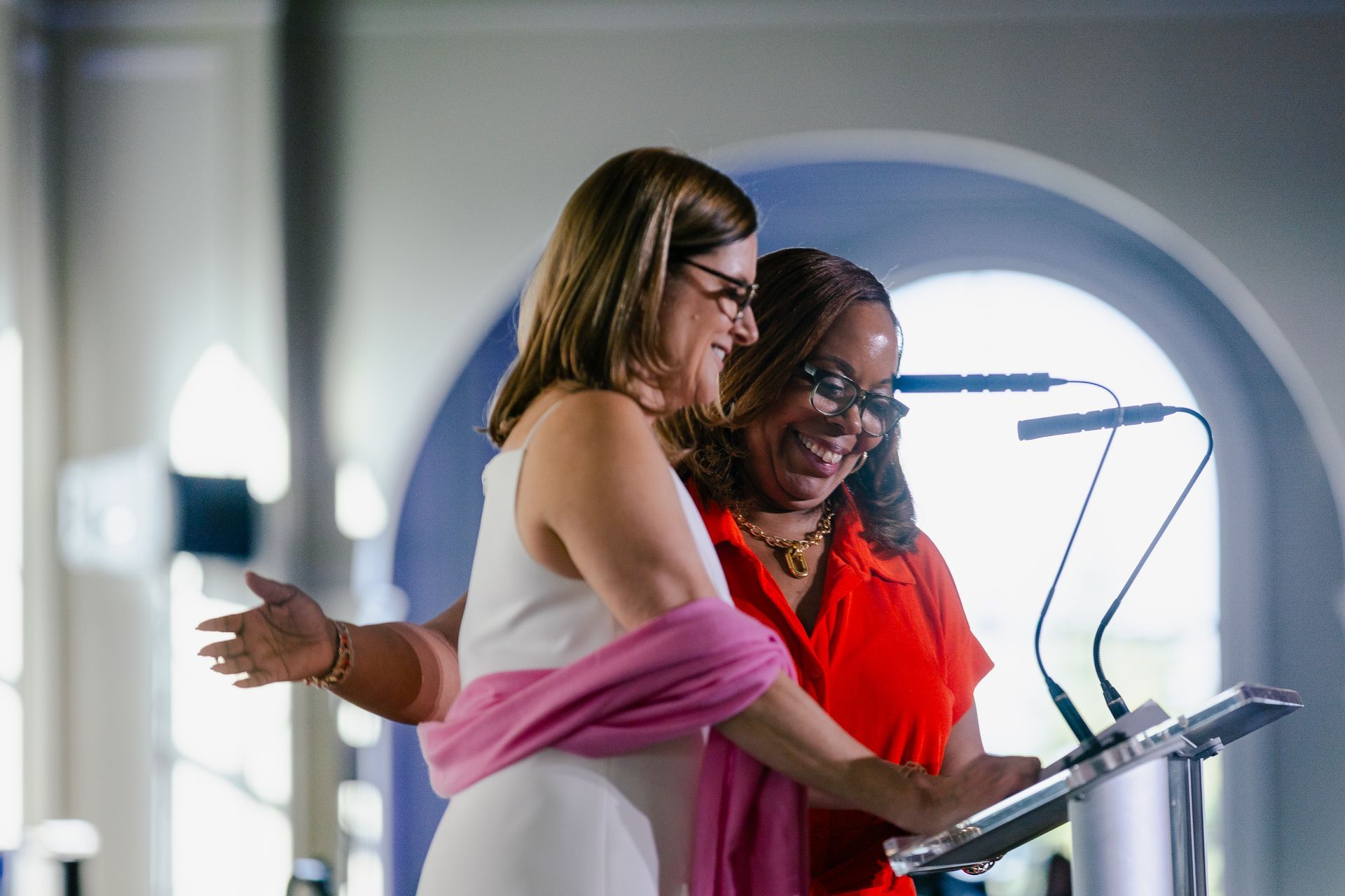 Two women at a podium, smiling and speaking during a presentation