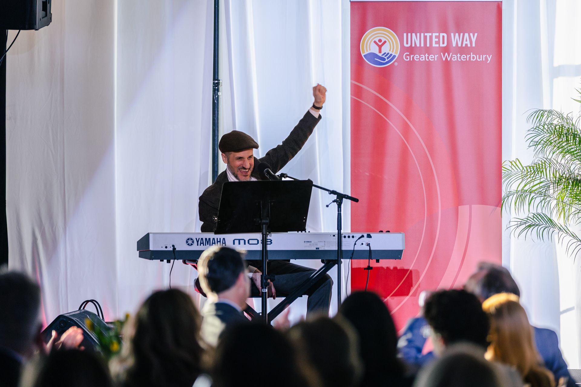 Musician at a keyboard onstage, raising one fist before a United Way banner, with audience in foreground