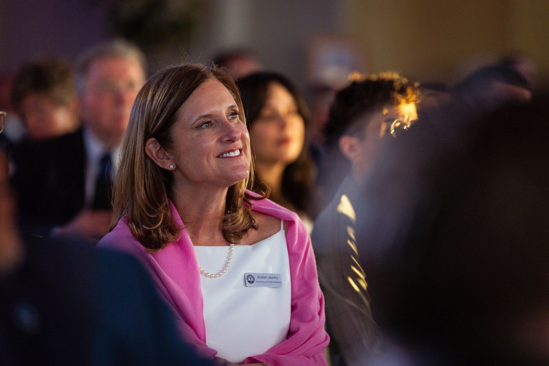 Woman in pink blazer smiling at an indoor event with people blurred in the background
