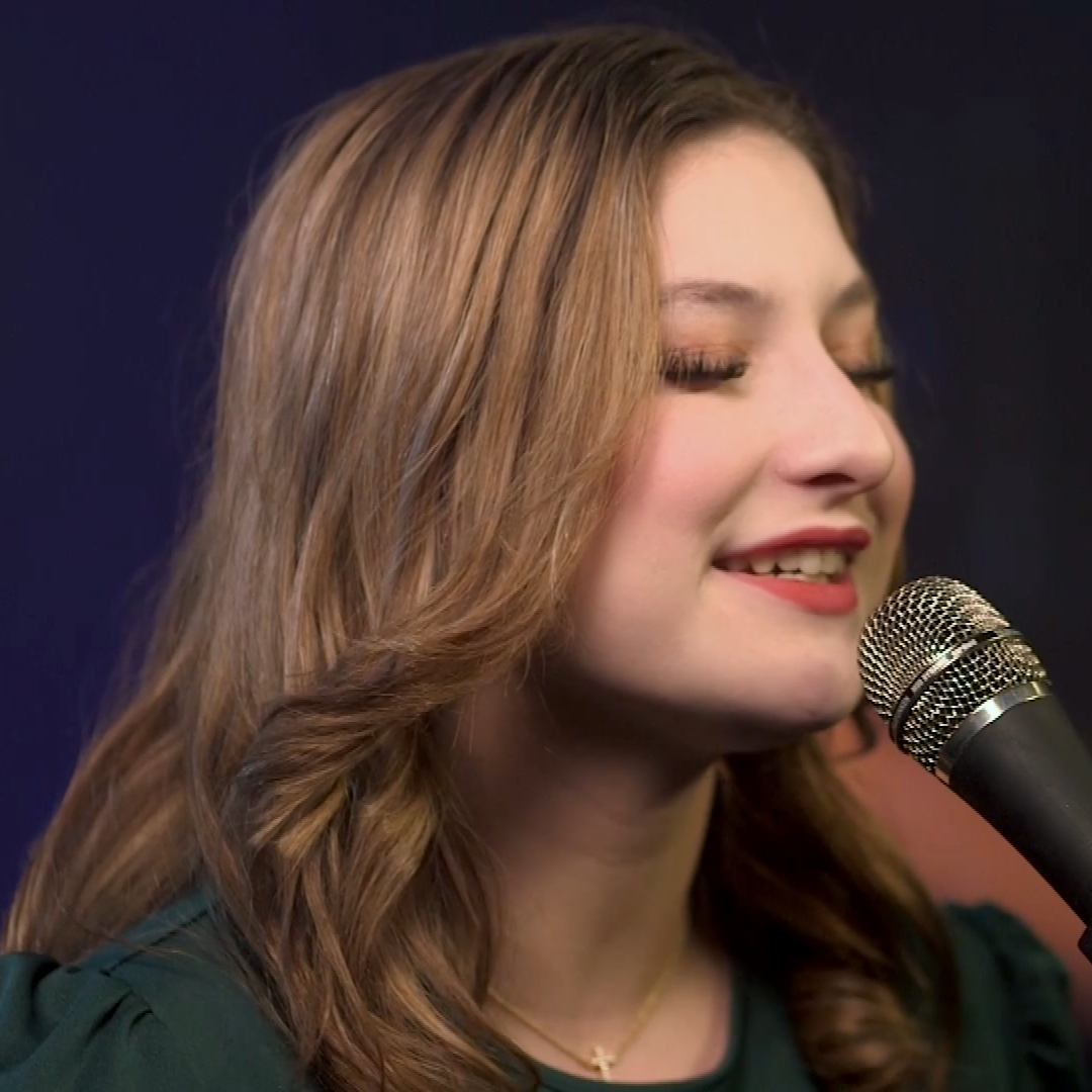Woman singing into a microphone on stage, eyes closed, with long brown hair and a dark background