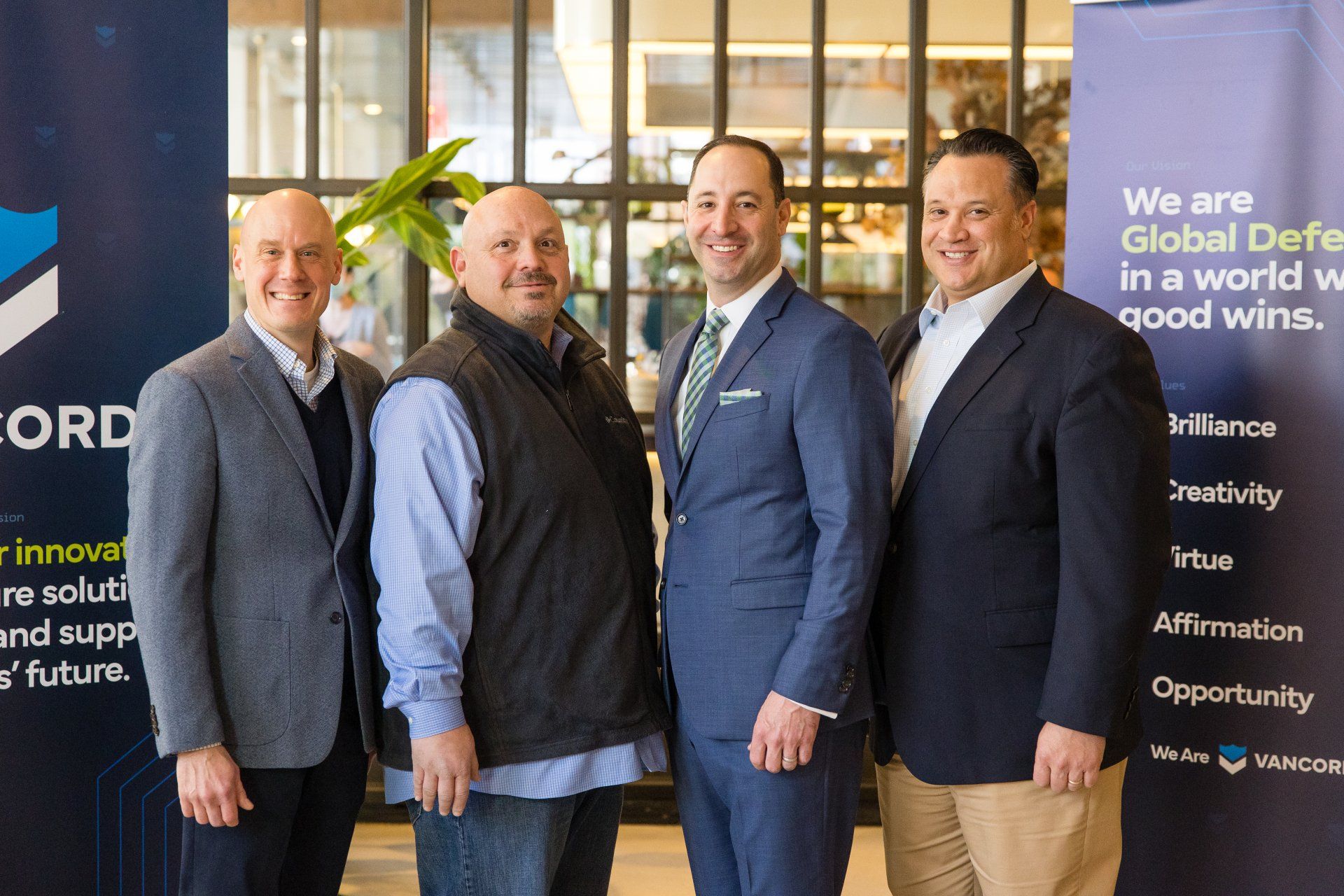 Four men in blazers pose indoors beside a conference banner about global defense.