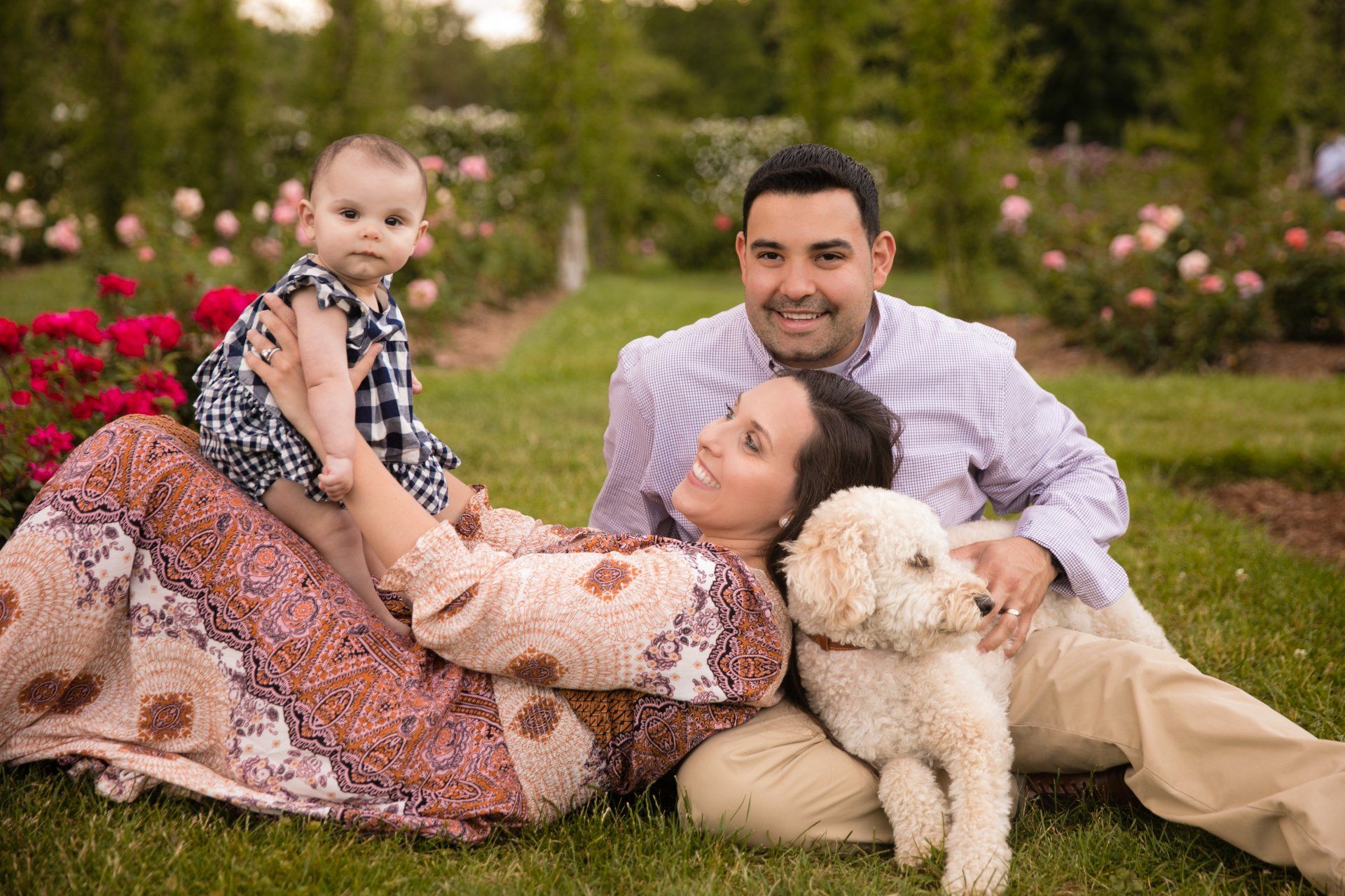 Family lying on grass in a garden, smiling with a baby and a small white dog.
