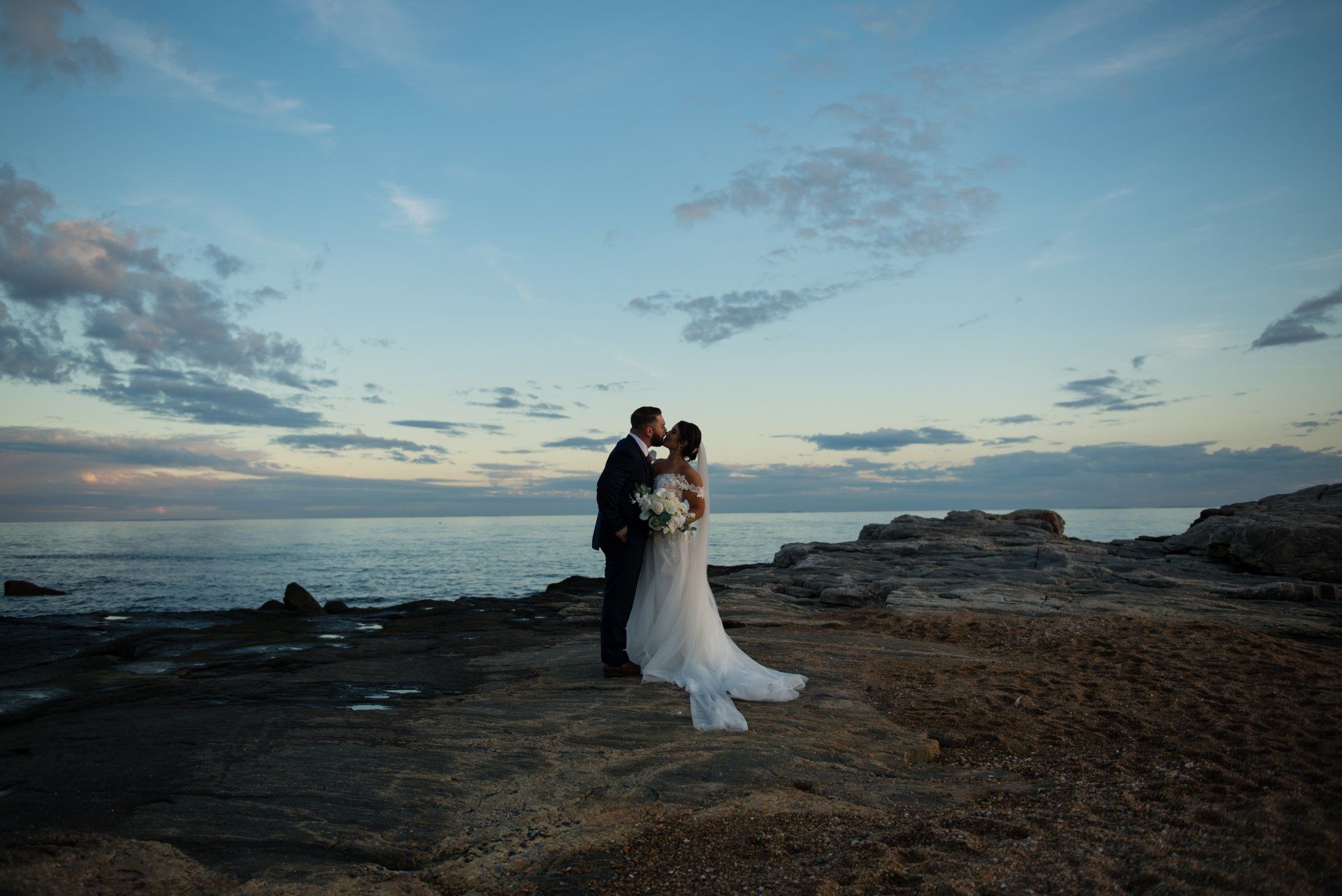 Couple in wedding attire embracing on a rocky beach at sunset under a cloudy sky