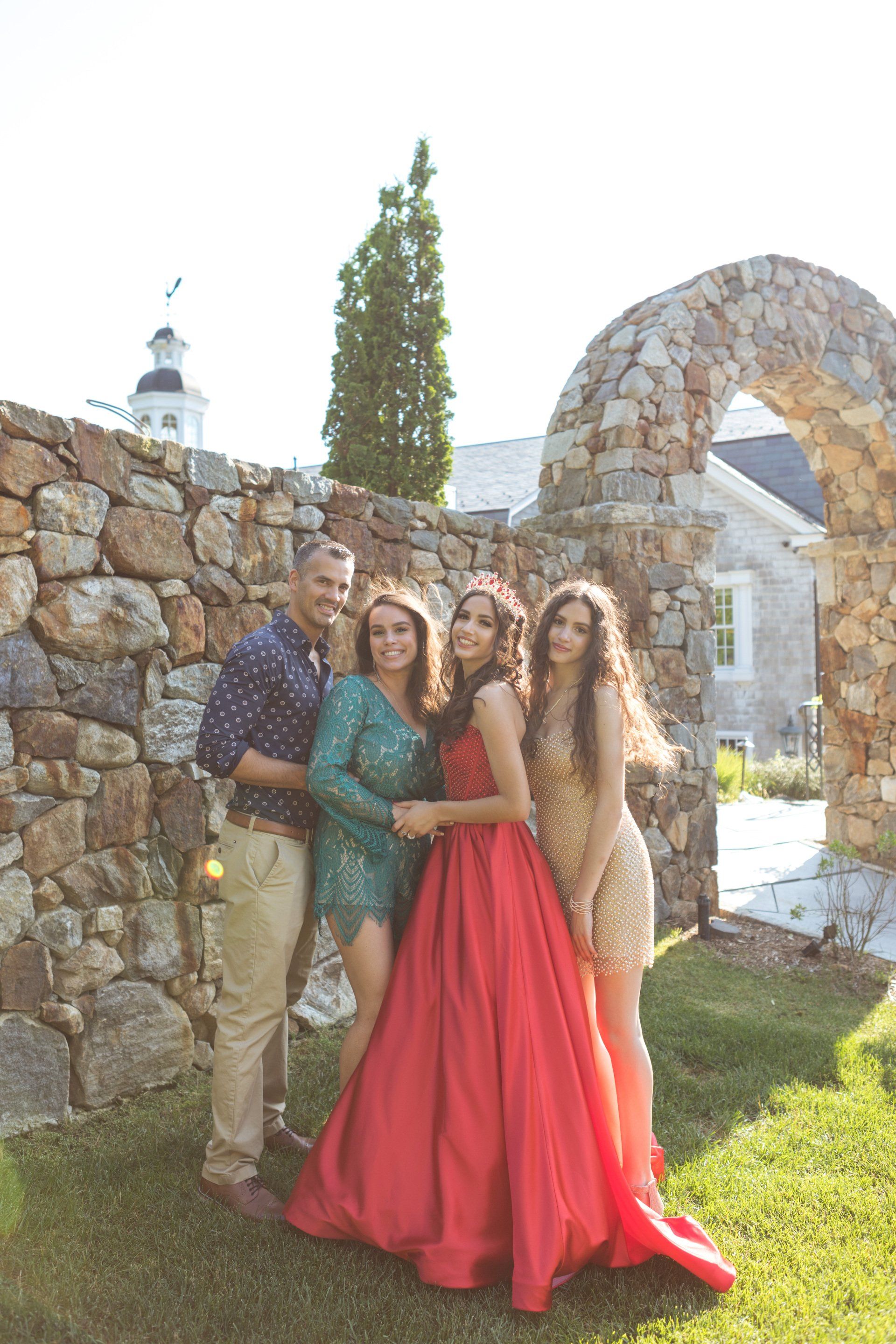 Four people posing outdoors by a stone wall and arch, one in a bright red dress.