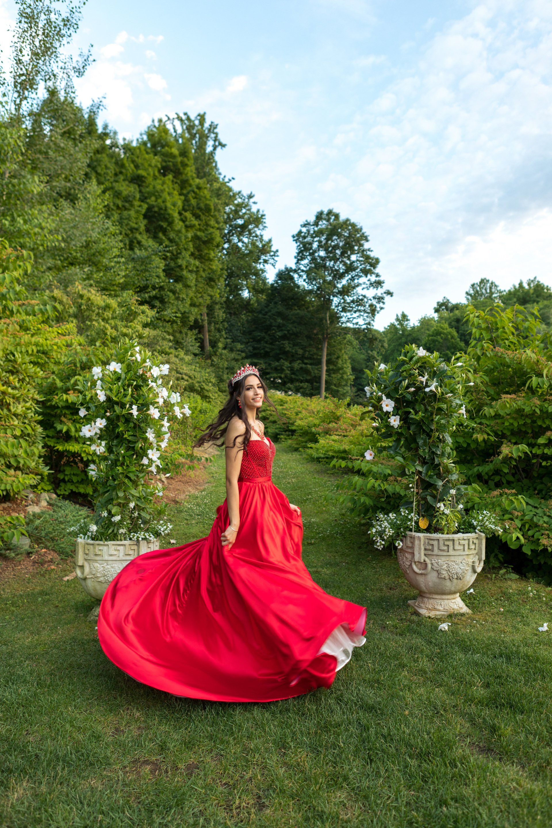 Woman in a flowing red dress twirls in a green garden with flowering bushes and stone planters.