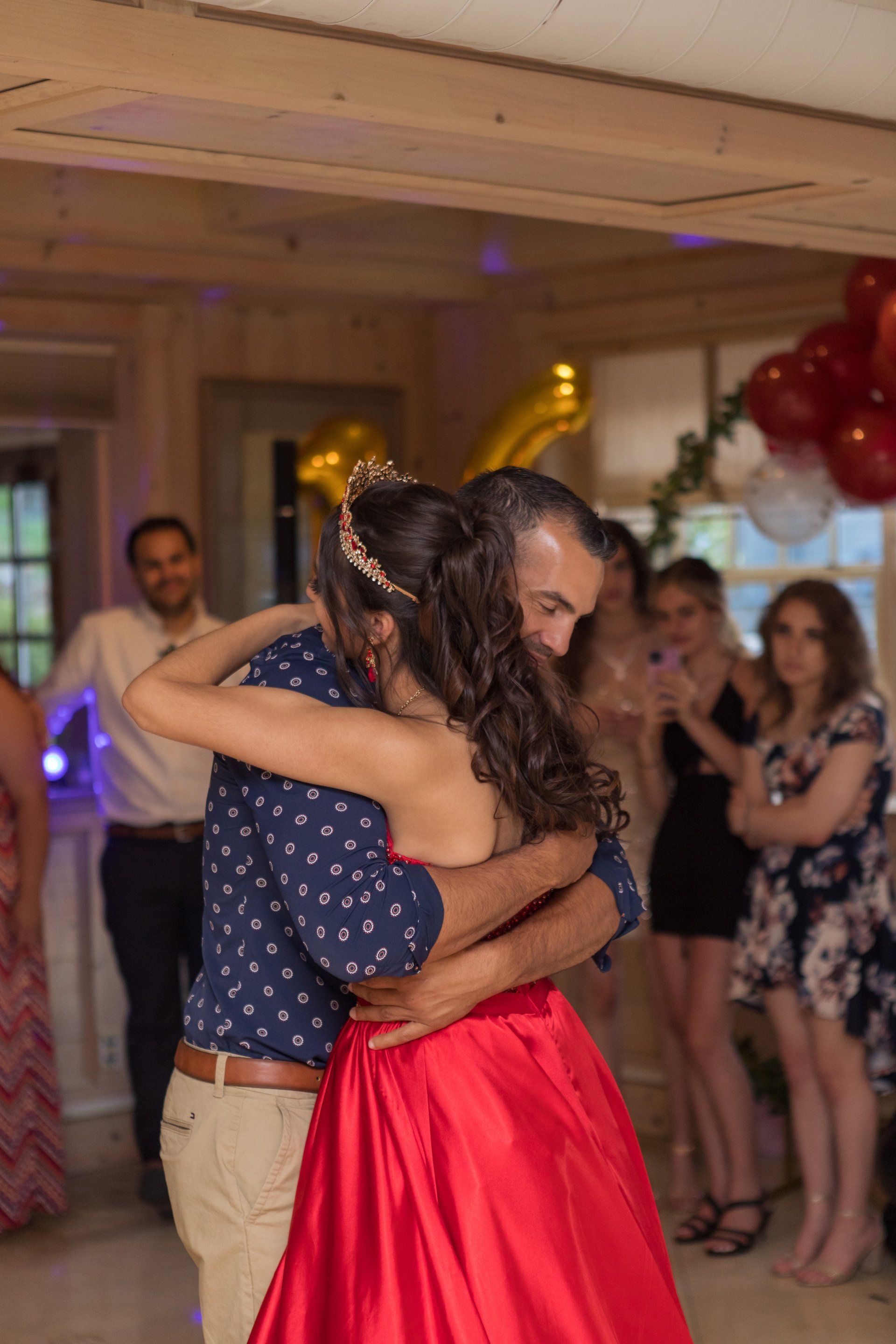 Two people embrace and dance at an indoor party, with guests and red balloons in the background.