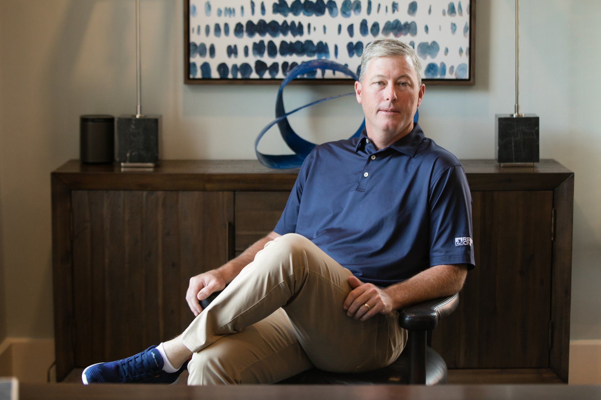 Man sitting in a chair in an office, wearing a navy polo and beige pants, with framed art and speakers behind him