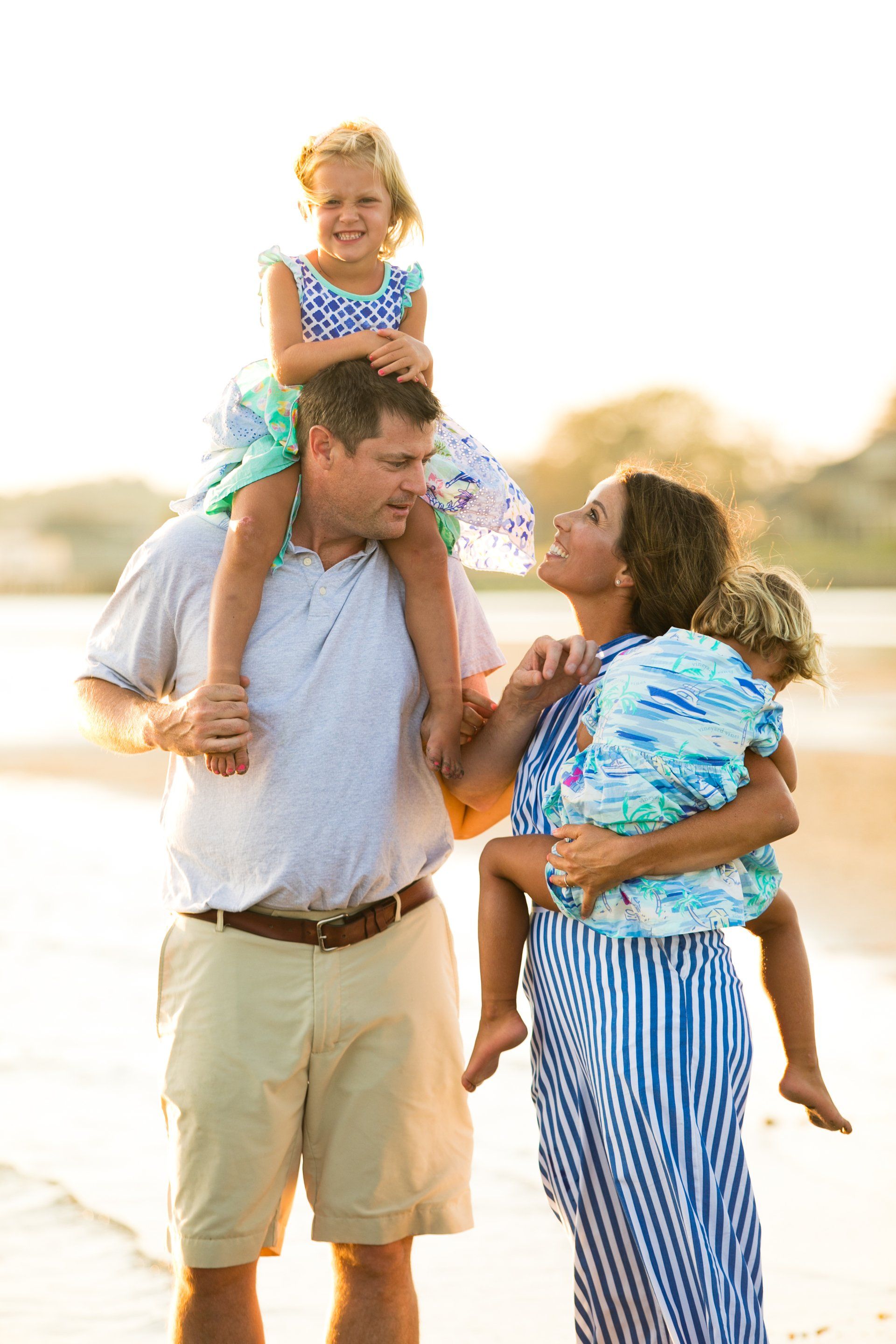 Family of four on a beach, smiling and carrying children on their shoulders near the water