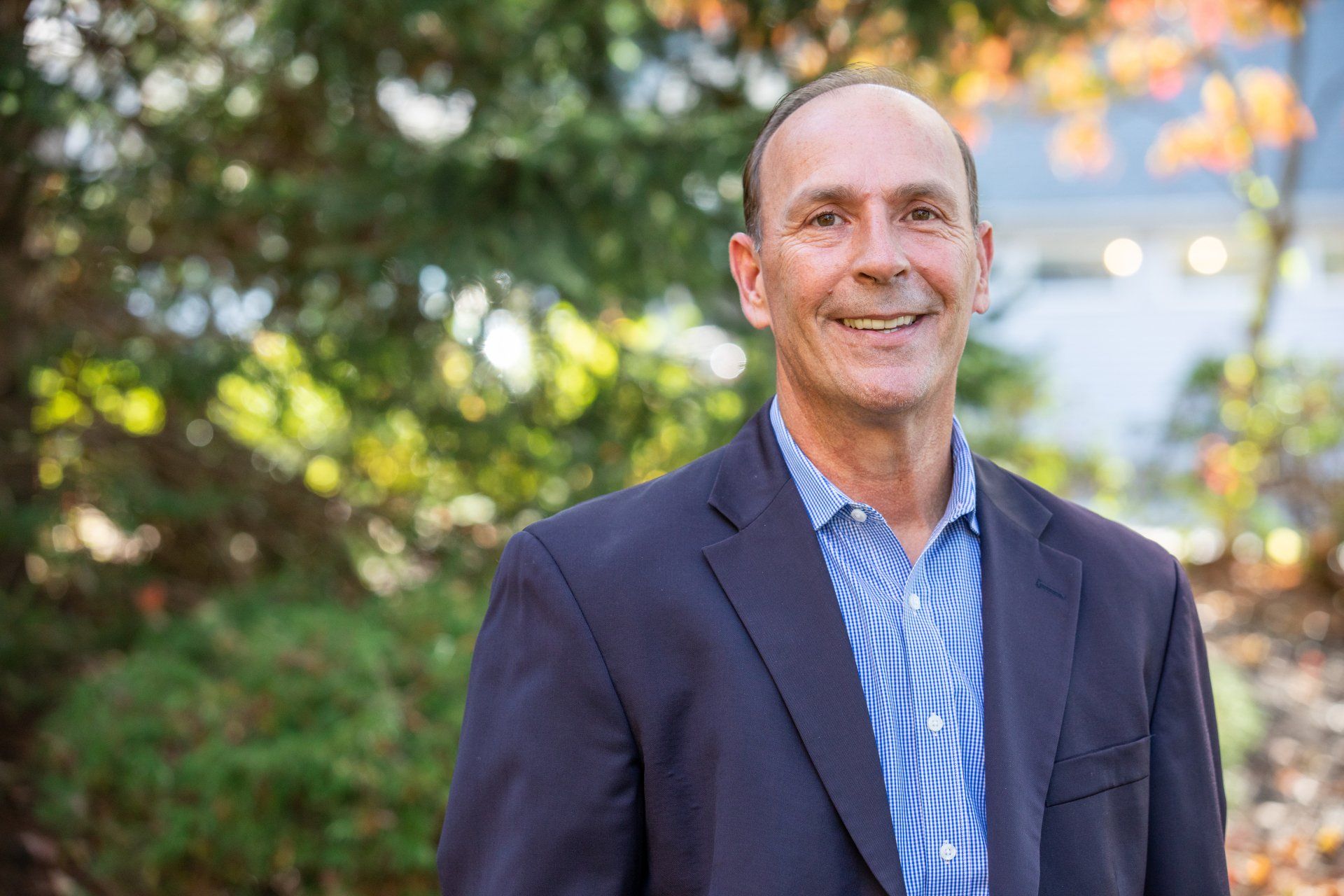 Man in a navy blazer and blue patterned shirt smiling outdoors with greenery and warm sunlight behind him