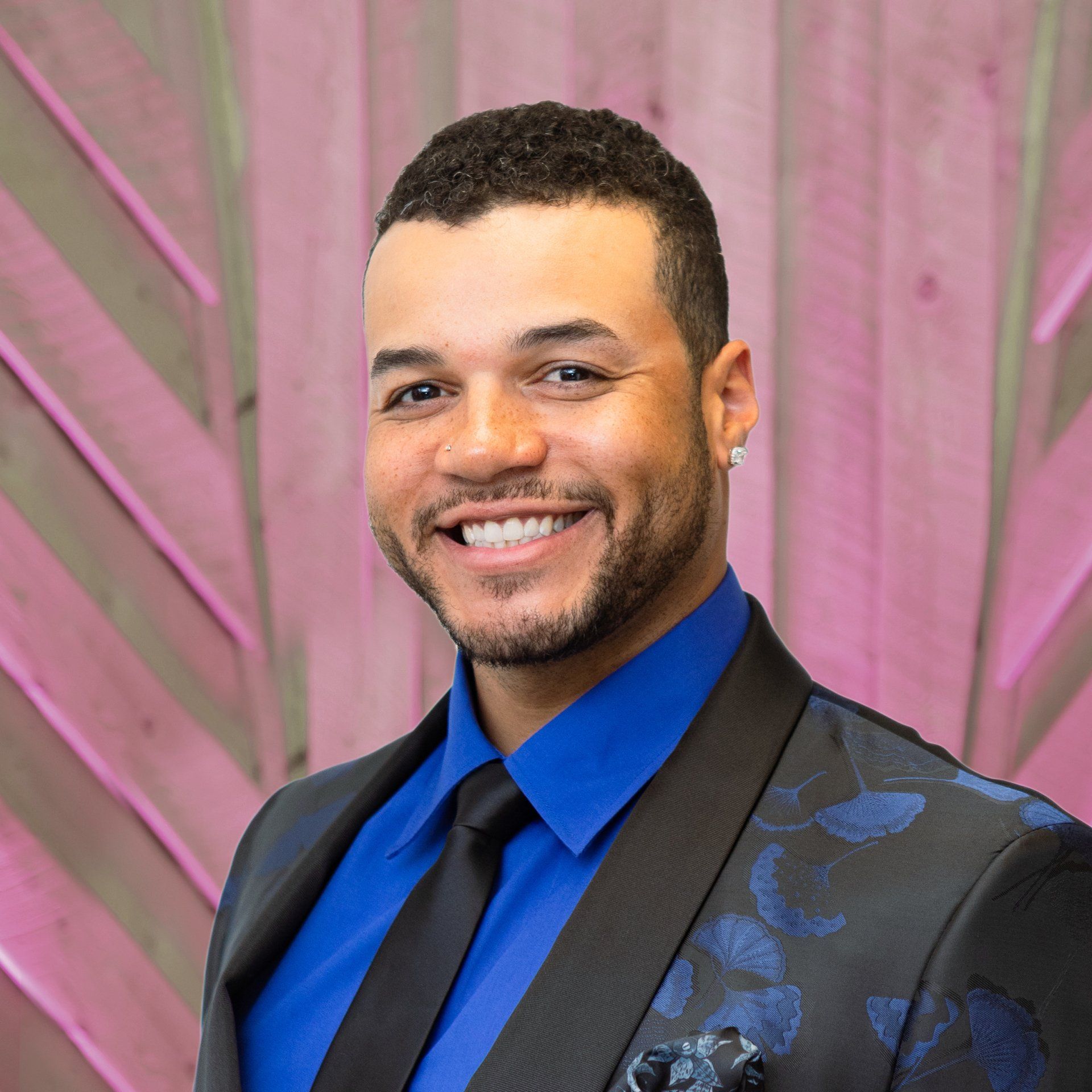 Smiling man in a blue shirt and black suit jacket against a pink draped backdrop