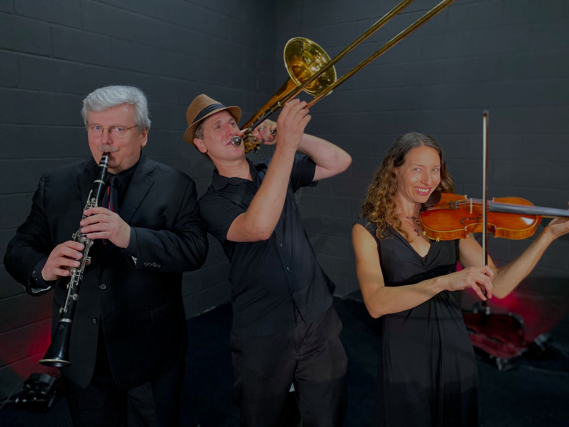 Three musicians perform together: clarinetist, trombonist, and violinist on stage against a dark backdrop.