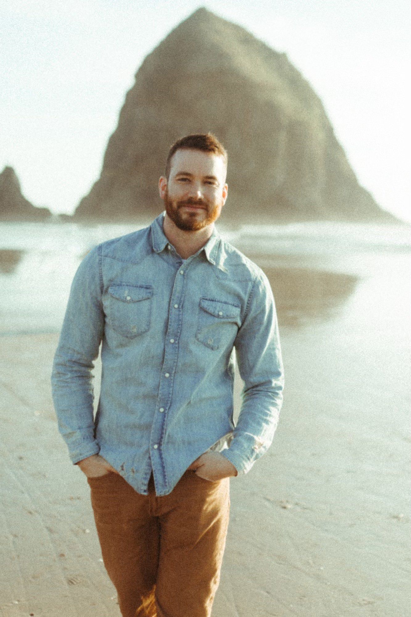 Man in denim shirt and brown pants standing on a beach with a large rock formation in the background