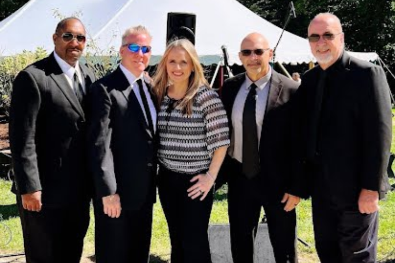 Five people posing outdoors in formal attire, standing together in sunlight.