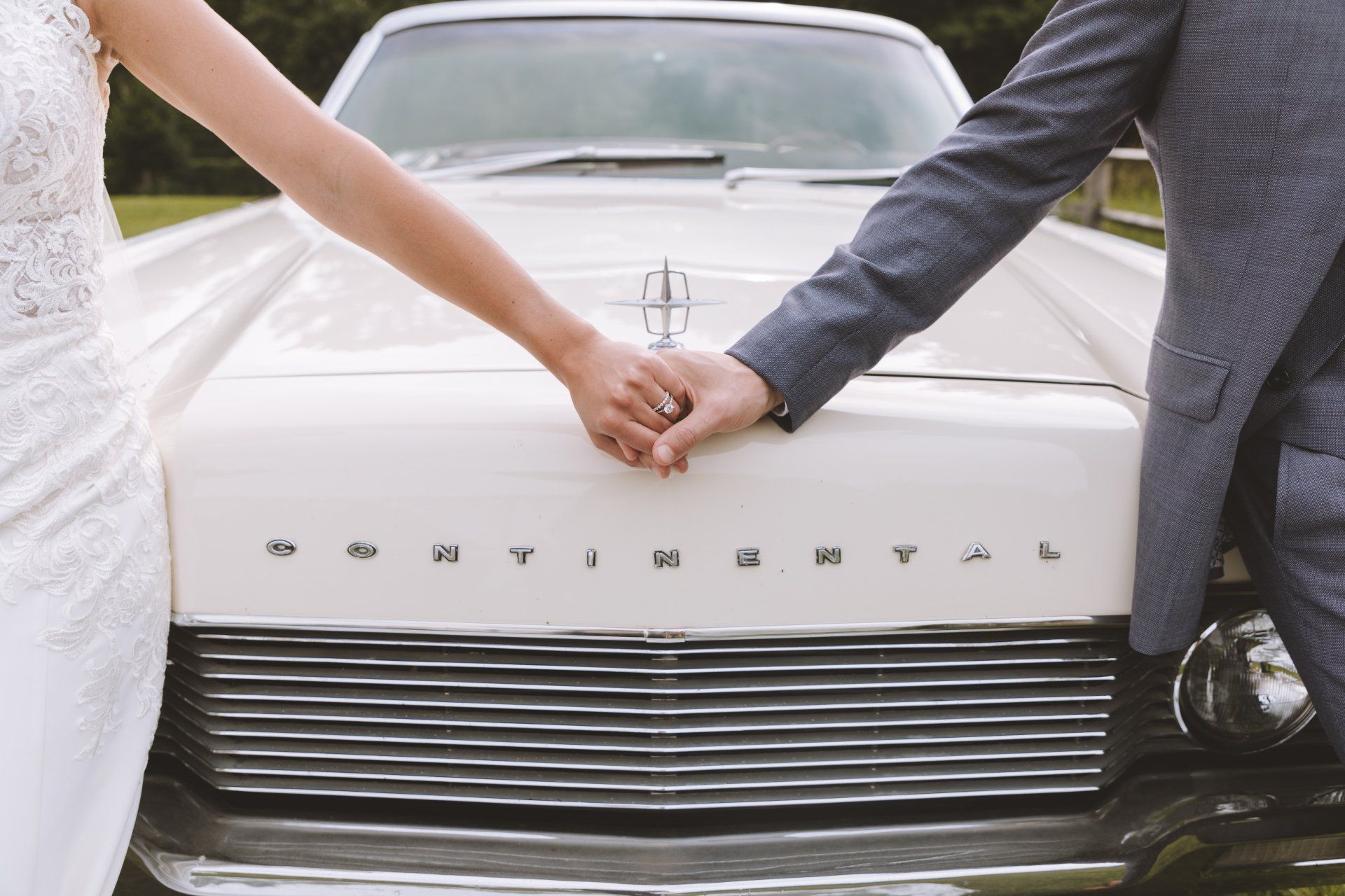 Bride and groom holding hands over a silver classic car hood
