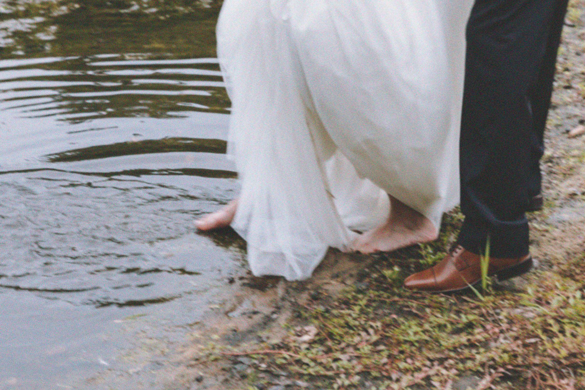 Couple standing barefoot in shallow water, the bride in a flowing white dress beside the groom in dark pants.