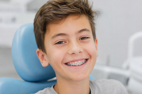 Bambino sorridente con l'apparecchio ortodontico seduto sulla poltrona del dentista, con un poggiatesta blu sullo sfondo.